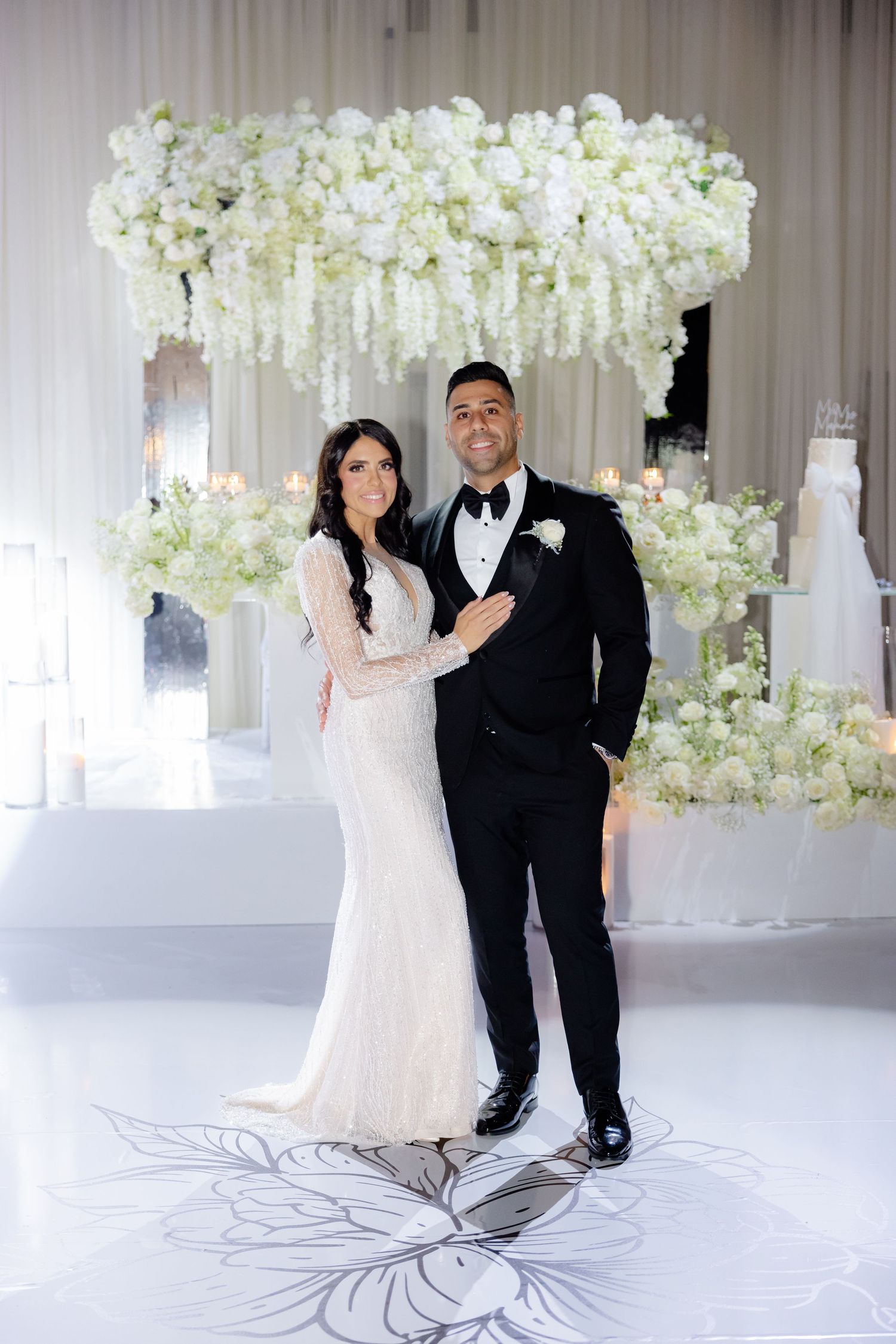Wedding couple standing beneath an elaborate white floral arch against a white backdrop with floor design.