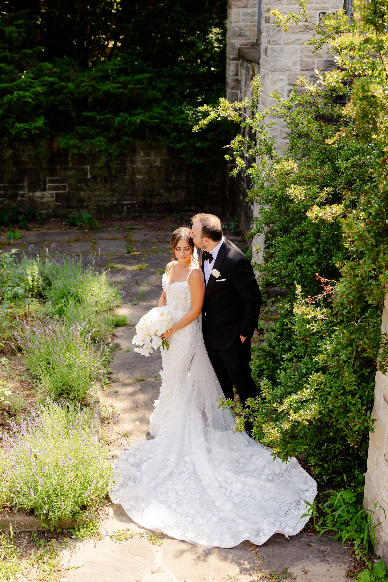 A romantic wedding couple embraces in a garden setting with a flowing white lace dress trailing on stone pathway.