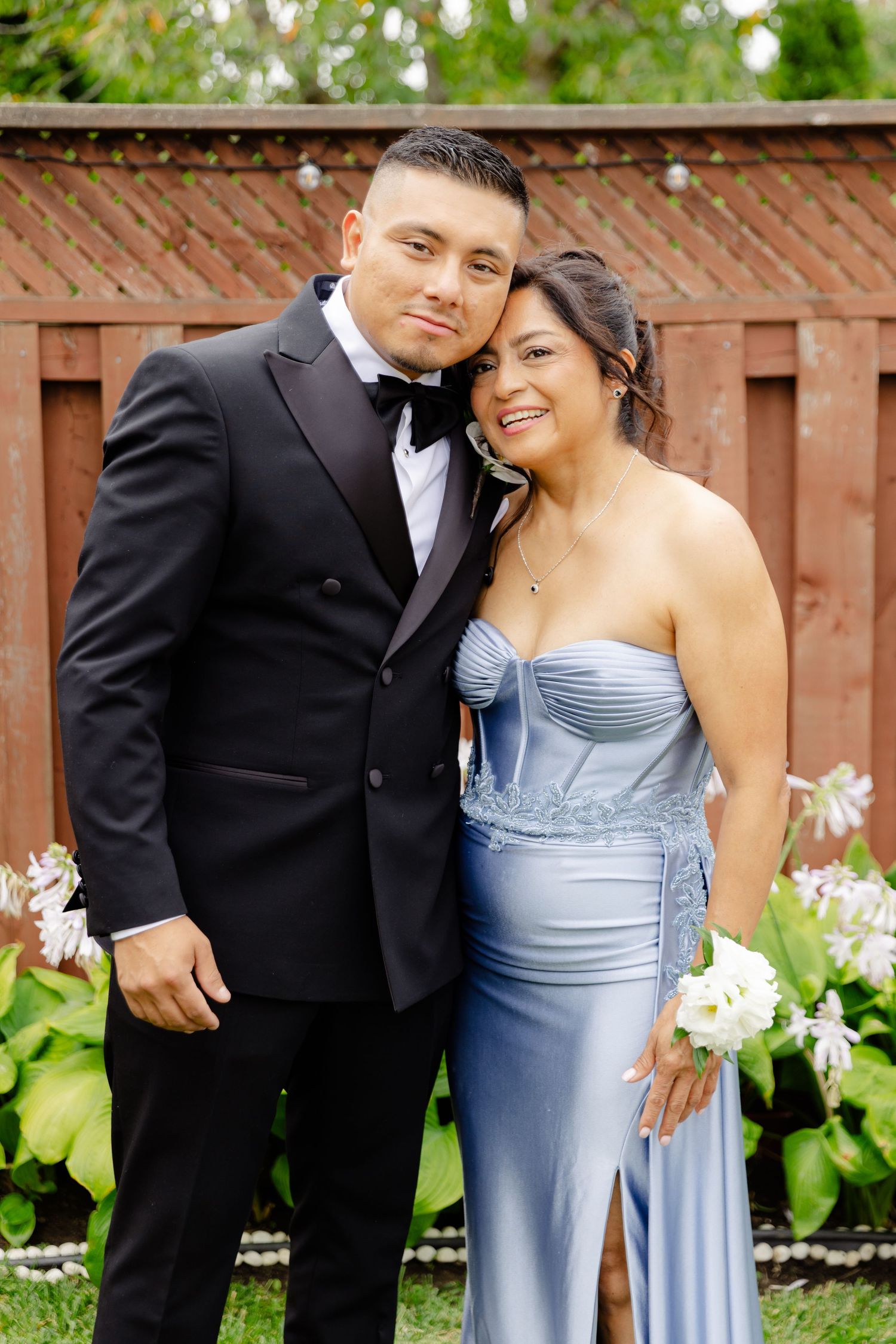 A couple in formal attire poses together against a garden backdrop with a wooden fence and white flowers in bloom.