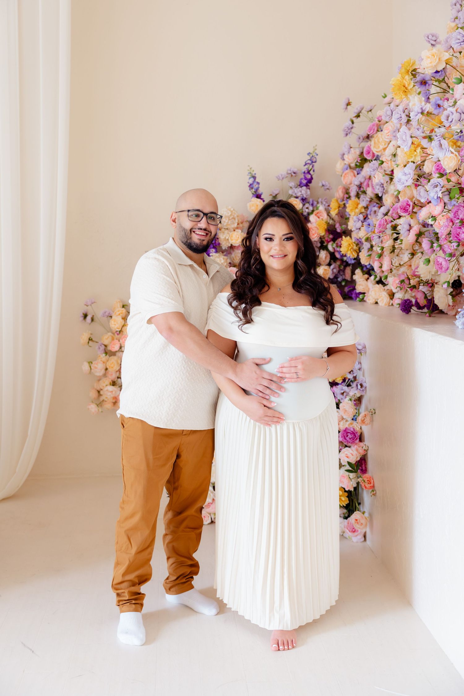 Couple in white outfits pose together by colorful floral arrangement during maternity photo shoot.