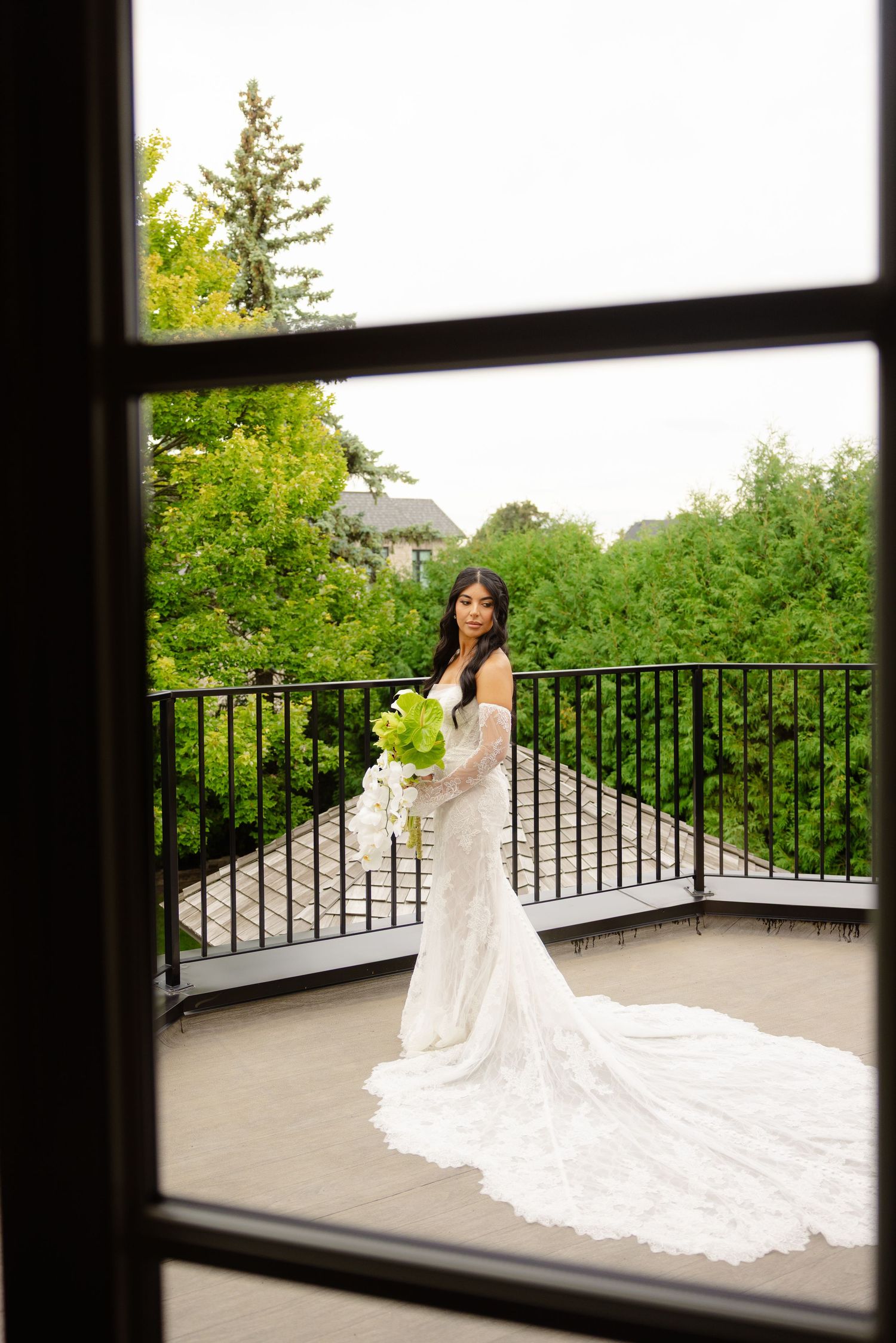 A stunning wedding dress with a long lace train is showcased on a balcony surrounded by lush green trees.