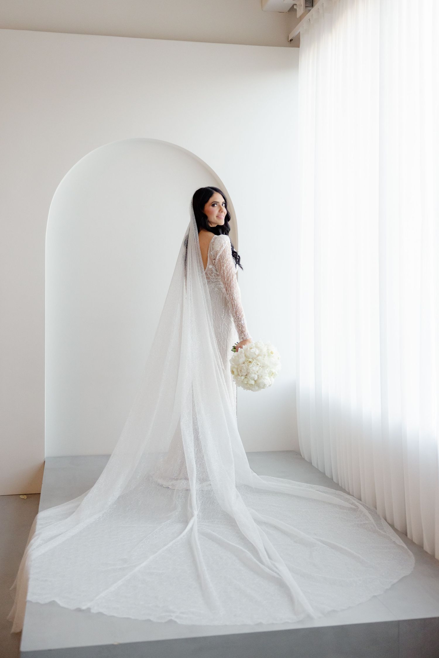 Bride with flowing veil poses gracefully in white architectural setting holding bouquet.