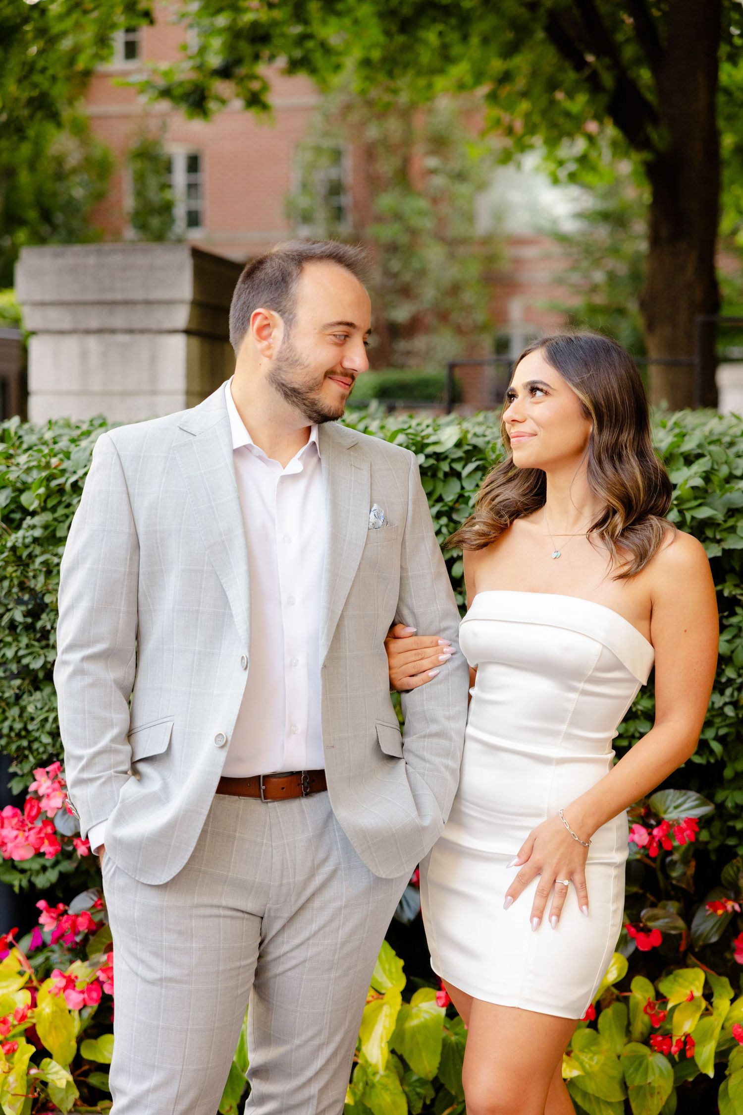 A couple shares a loving glance while posing in front of pink flowers and green hedges.