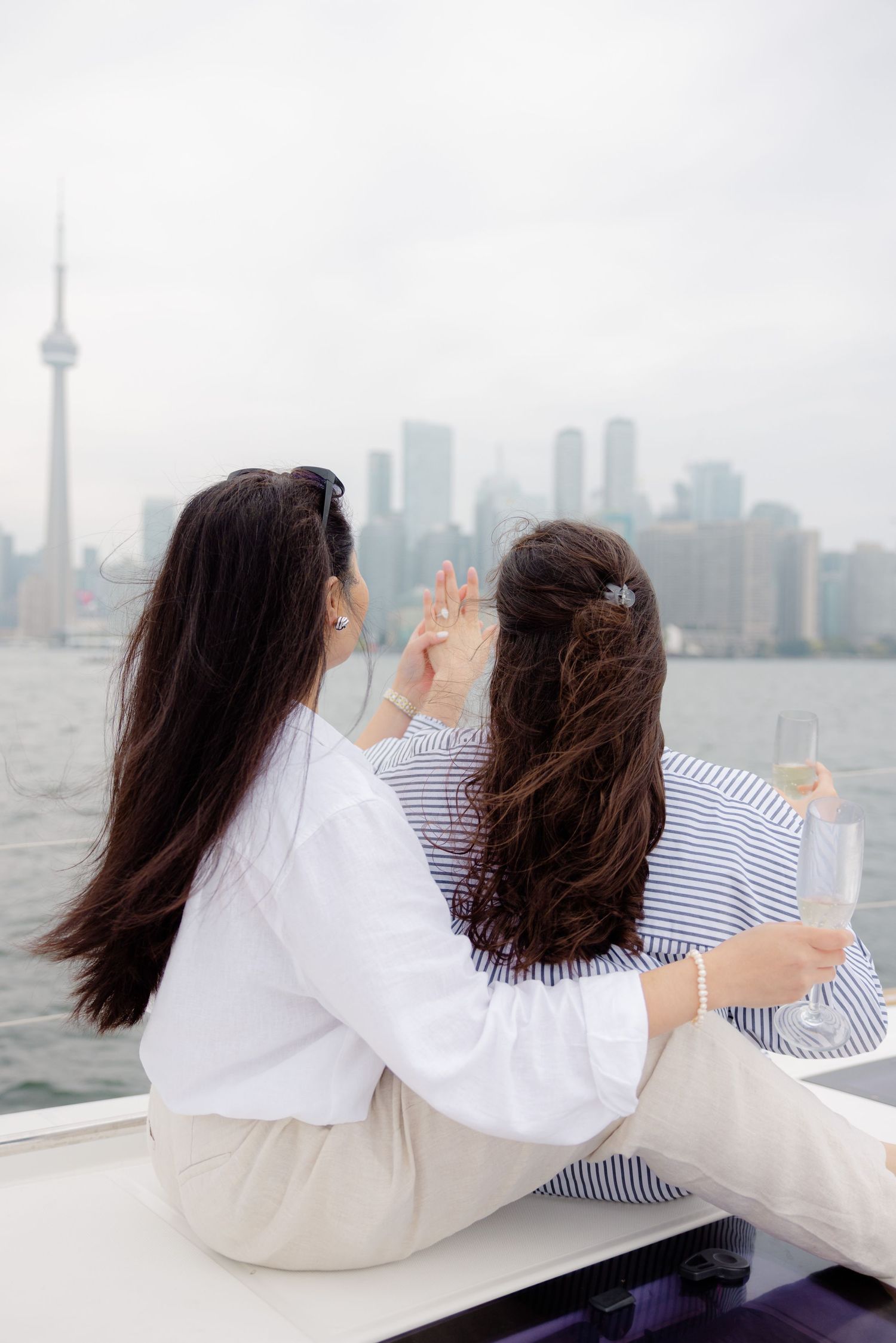 Two women with long dark hair sit together overlooking a cityscape with skyscrapers and a tall tower in the distance.