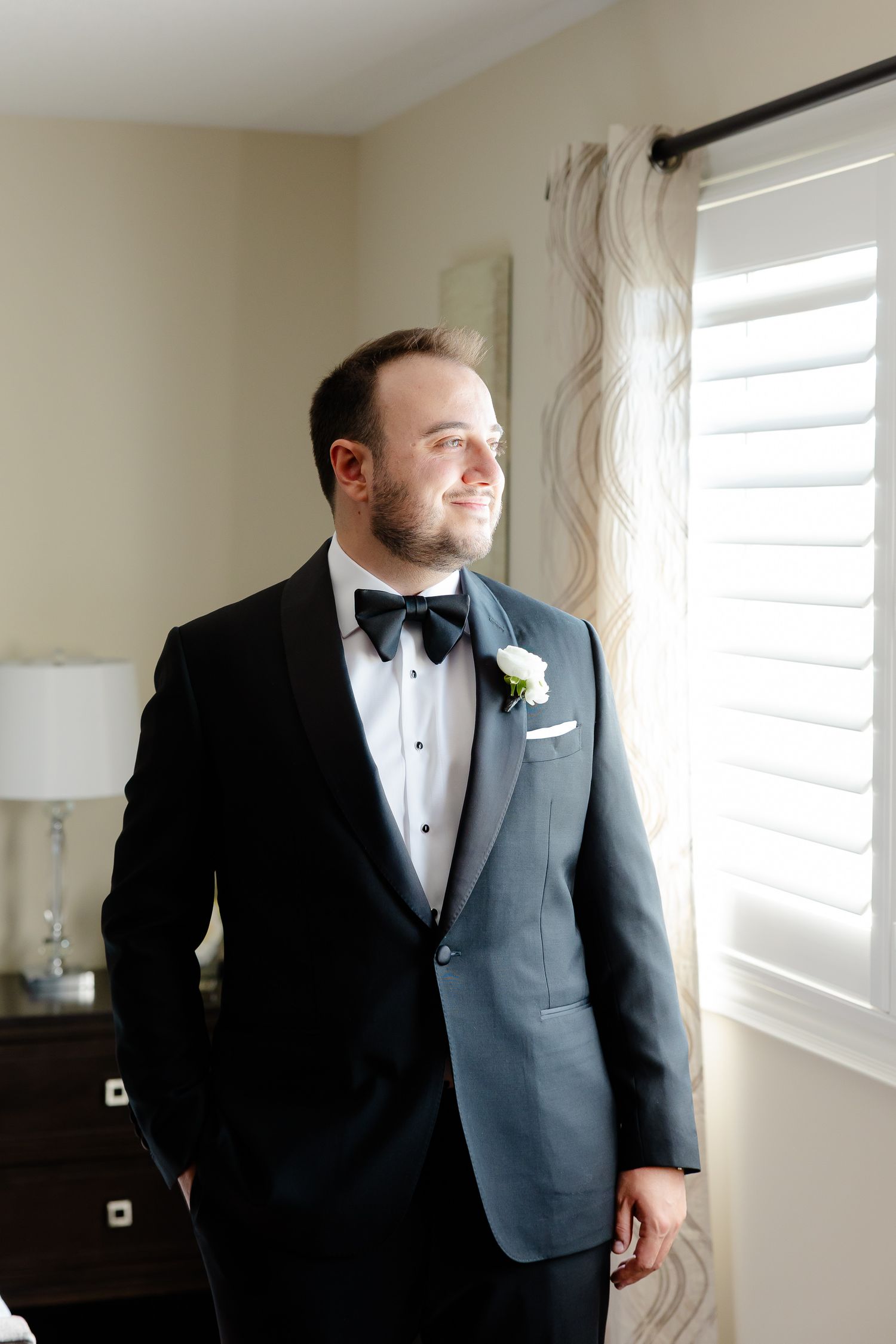 A series of four photos showing someone in a formal grey suit and black bow tie getting ready in a bright room with shutters.