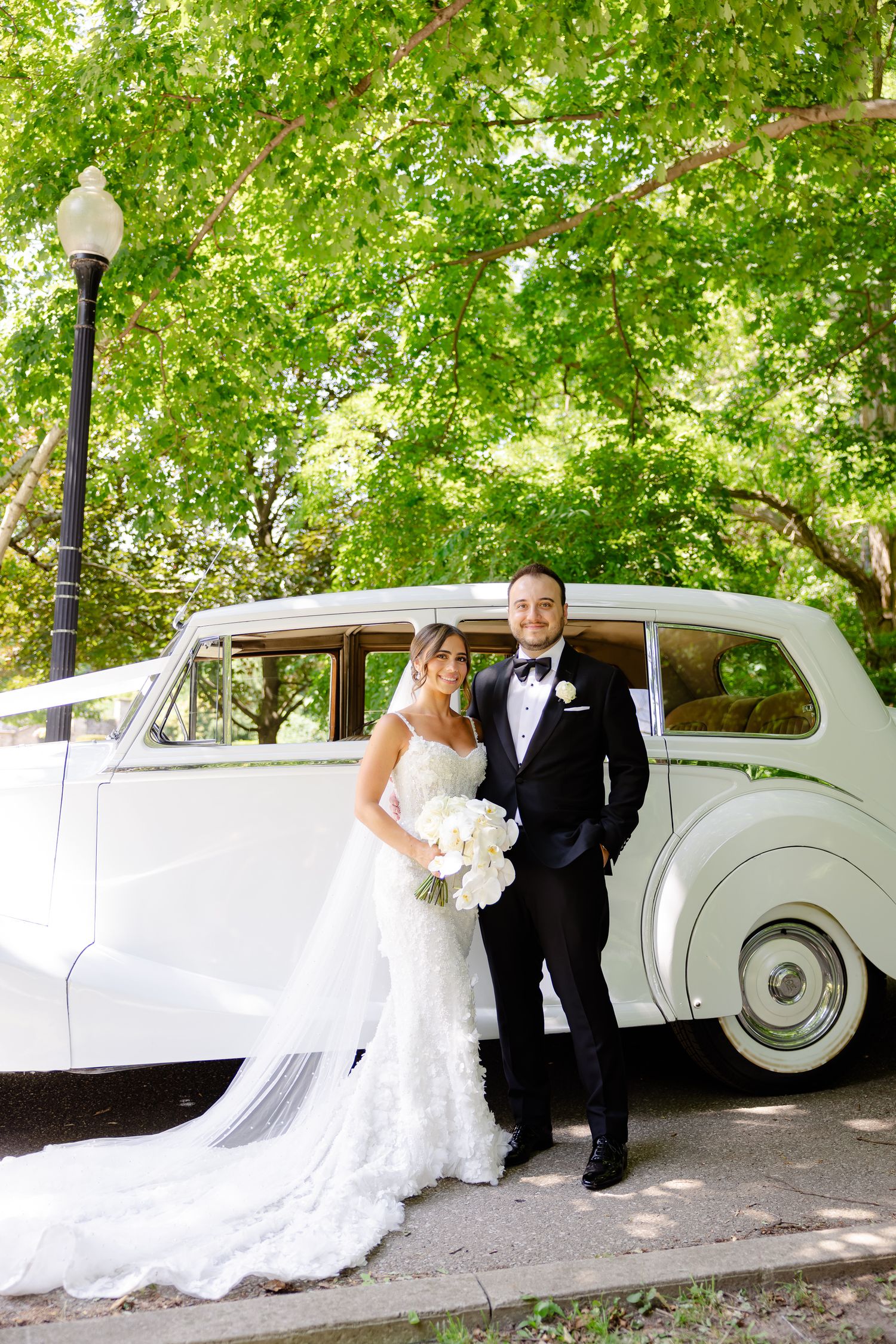 A classic white Rolls Royce provides an elegant backdrop for a couple's wedding day photo under lush green trees.