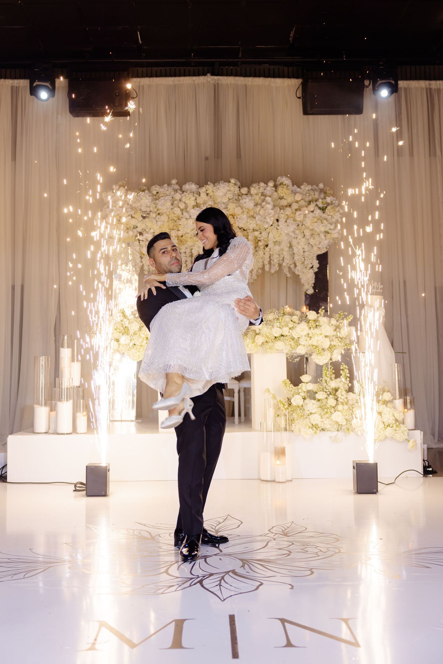 Romantic lift pose under floral arch with sparkler effects on personalized dance floor.