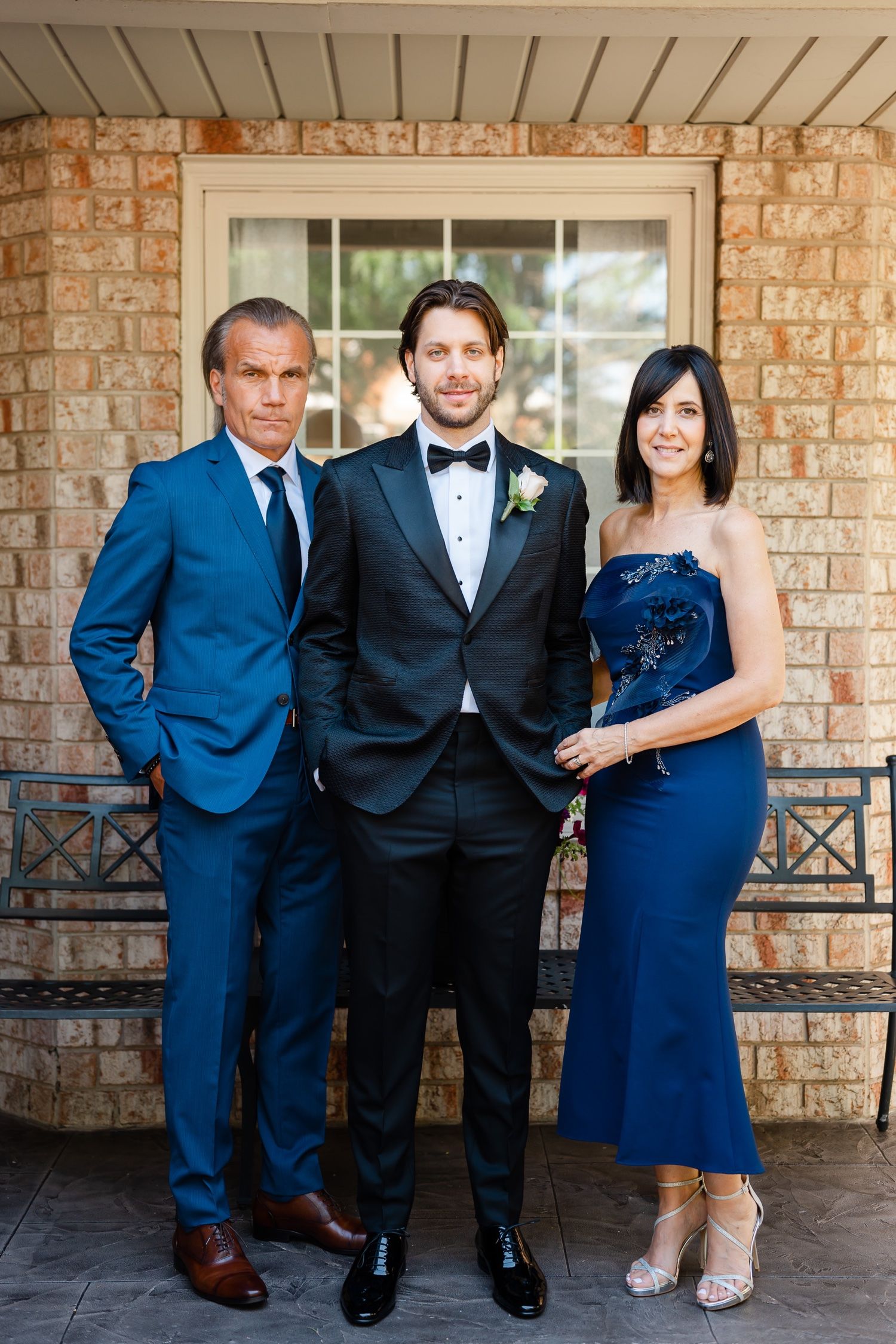 Wedding party portrait with three people in formal attire against stone house exterior.