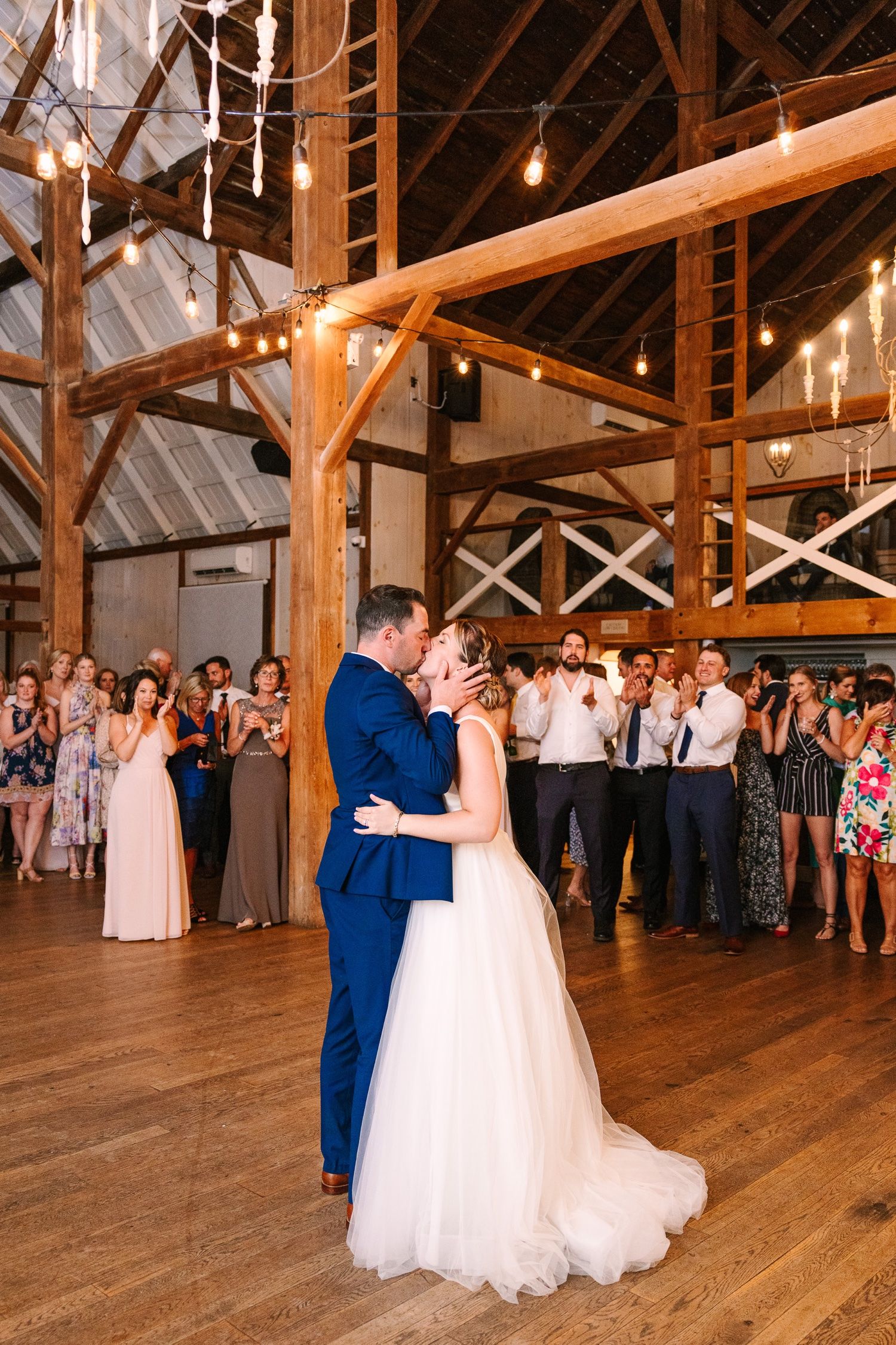 First dance surrounded by wedding guests in rustic barn venue with exposed wooden beams.