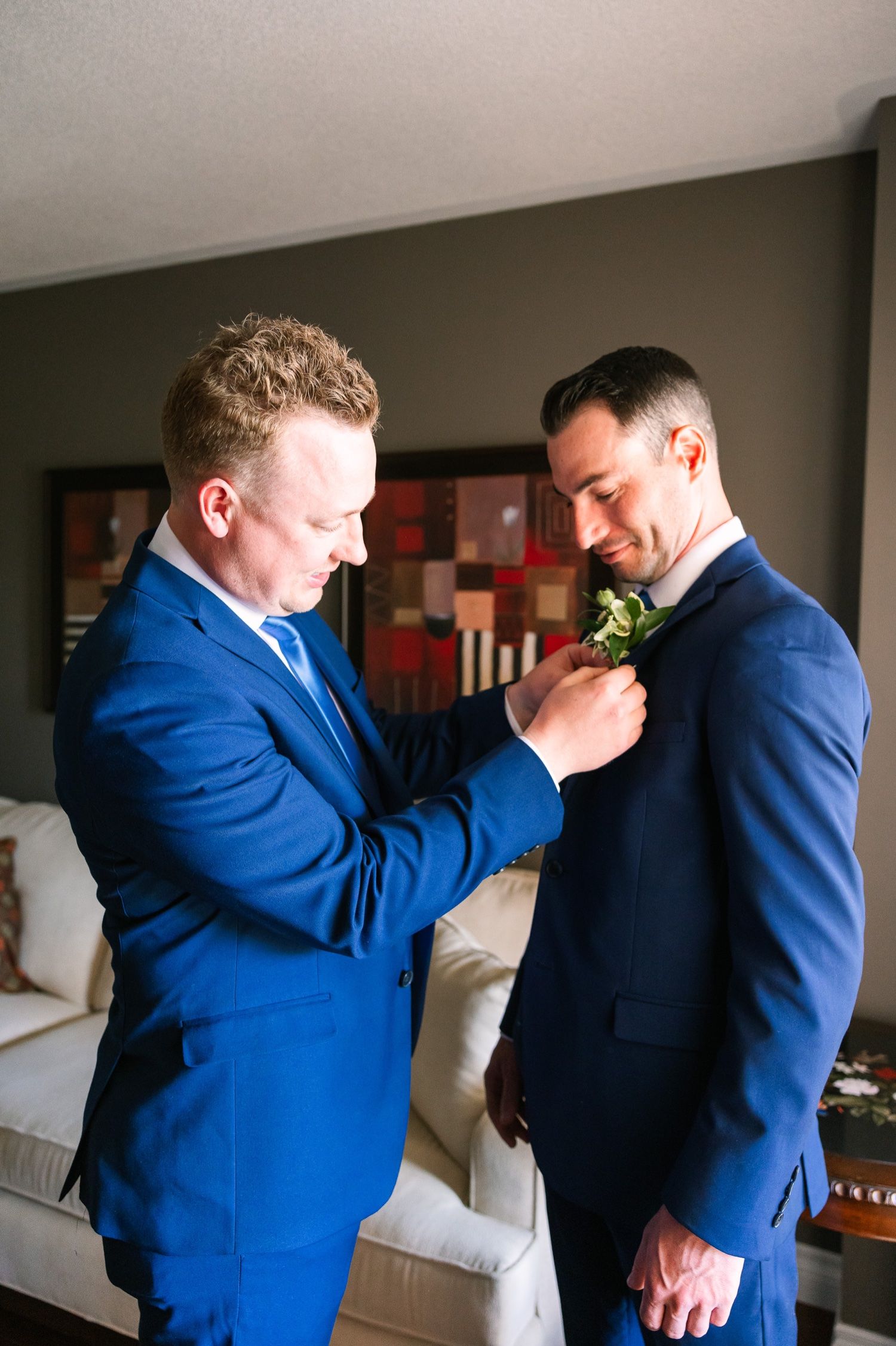 Two people in blue suits help each other with boutonnières before a formal event.
