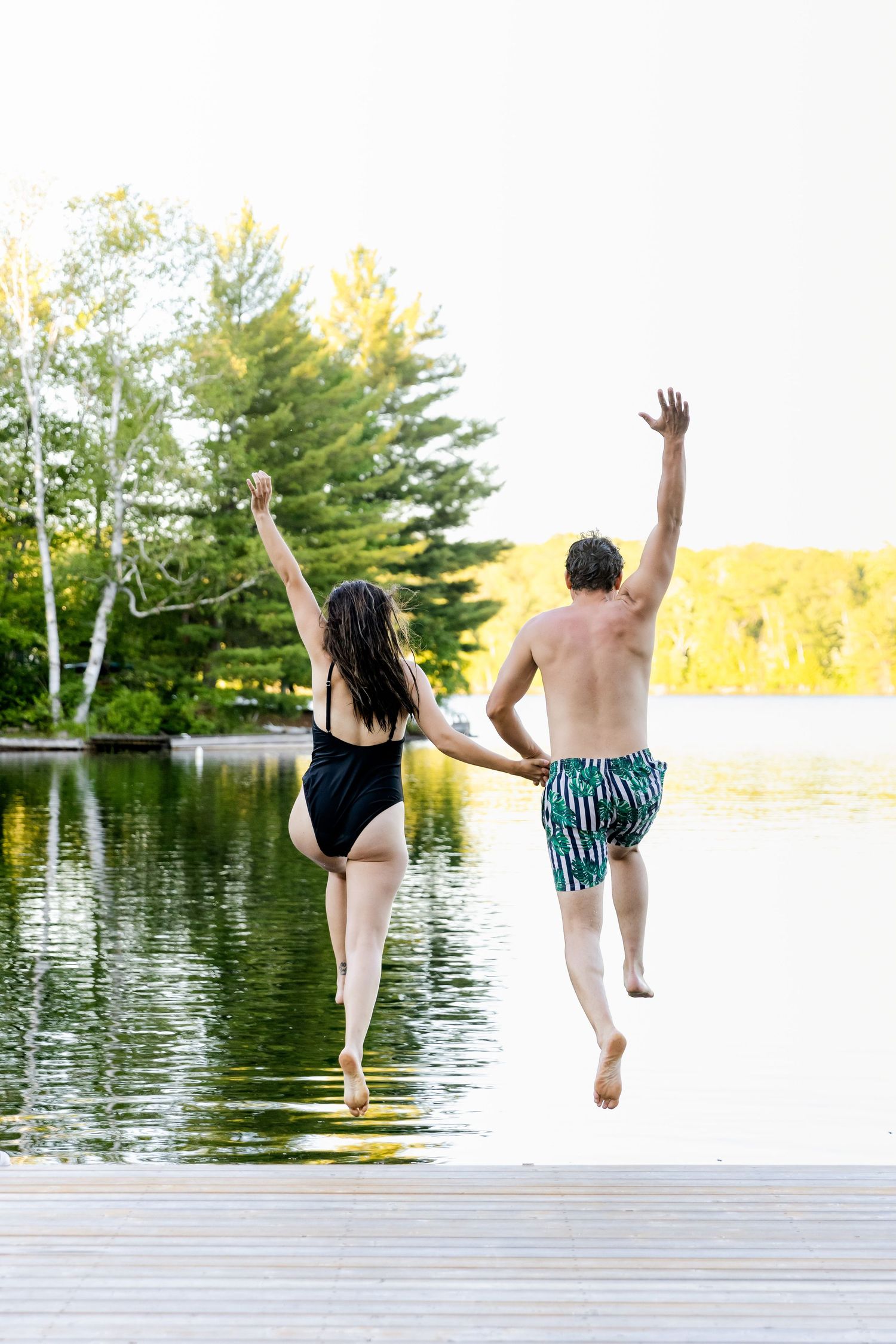Couple in swimwear joyfully jumping off a dock into a lake surrounded by pine trees on a sunny day.