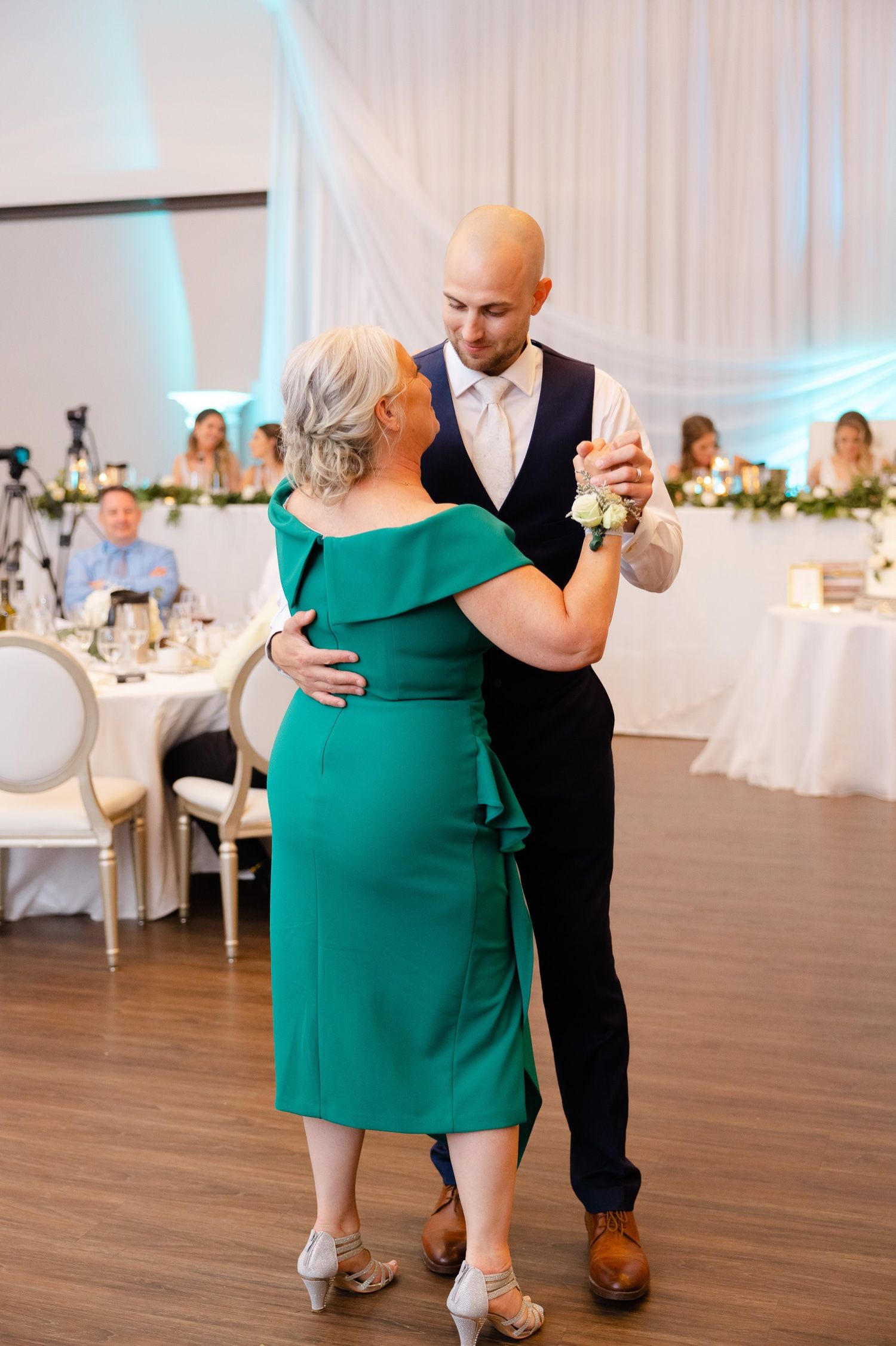 A couple dances together at a wedding reception as she wears an elegant emerald green dress and he wears a formal suit.