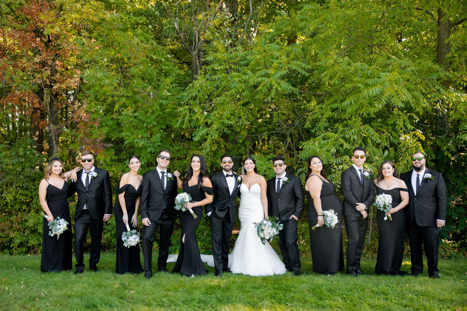Large wedding party in black formal wear poses together on grass with green trees in background.