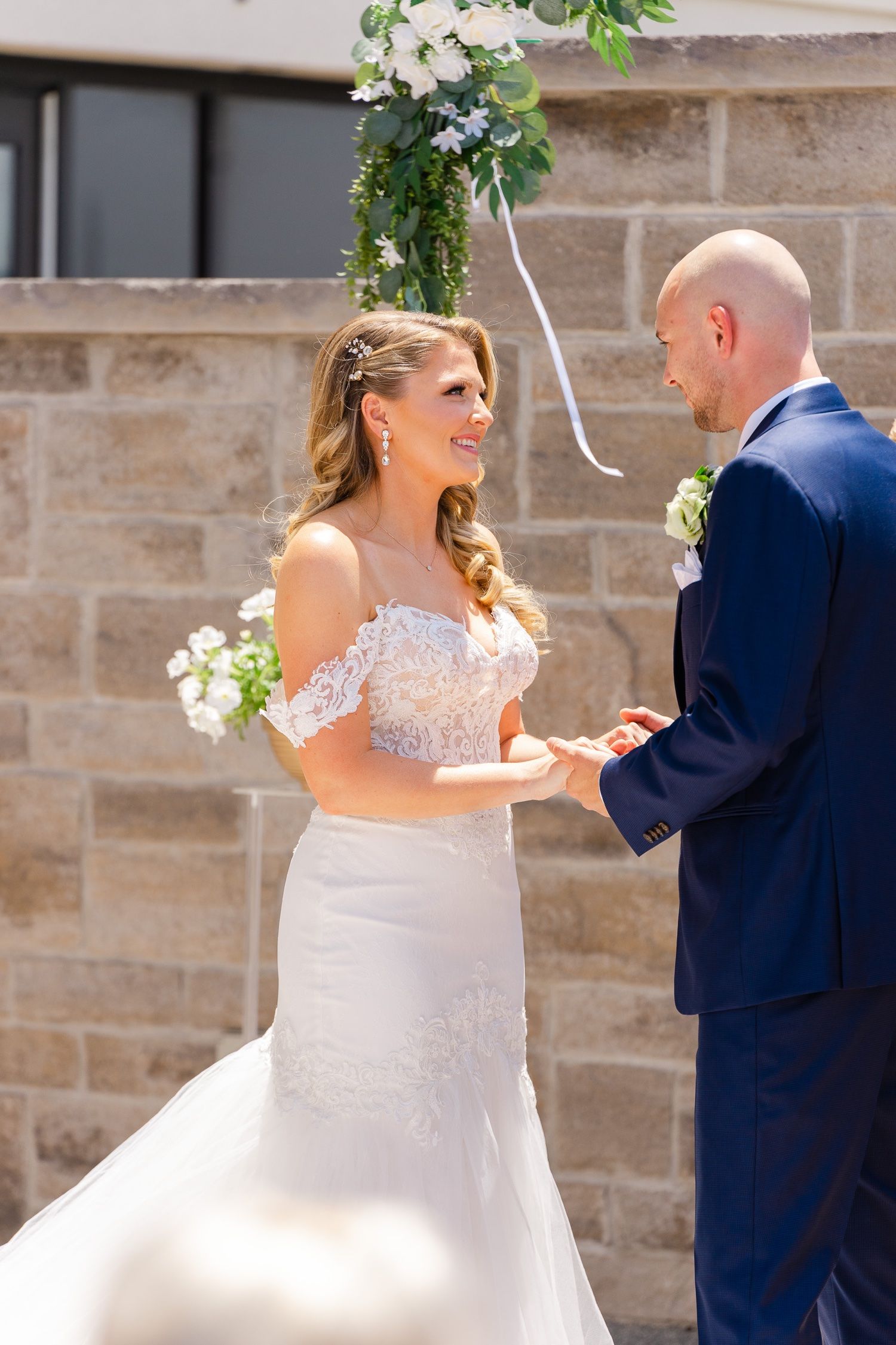 A beautiful outdoor wedding ceremony with a couple exchanging vows under floral decor against a stone wall backdrop.