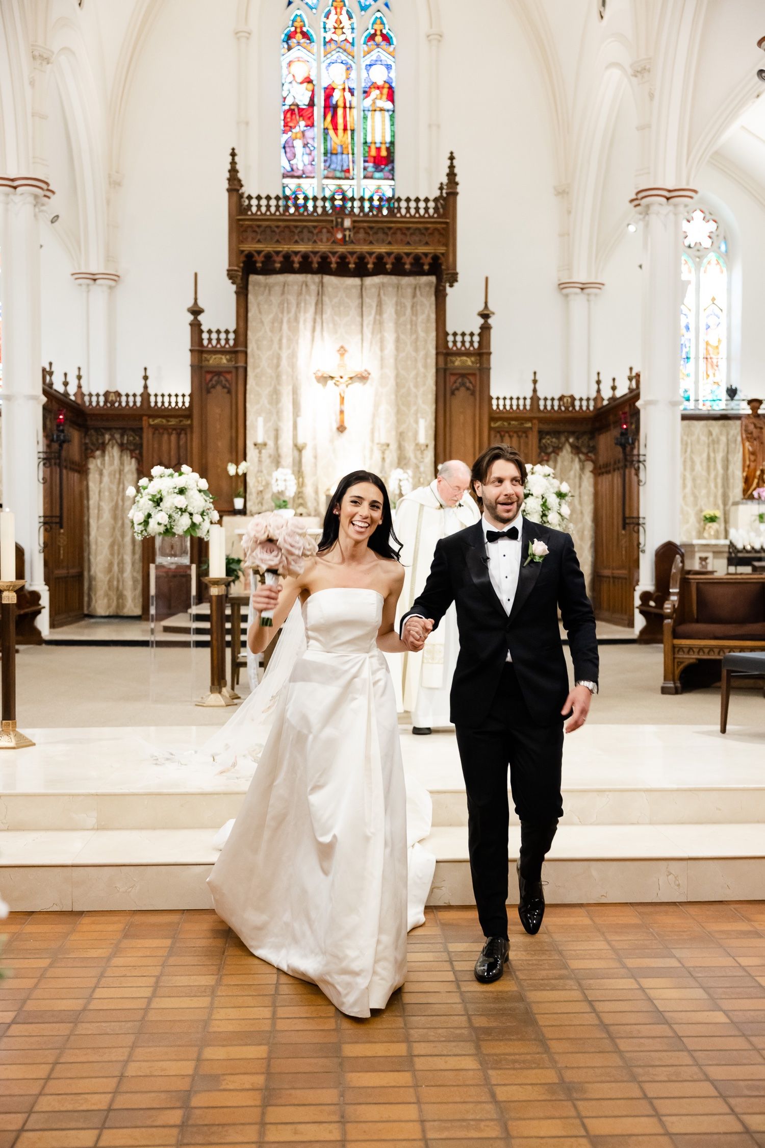 A couple walks down church aisle after their wedding ceremony with stained glass windows in background.