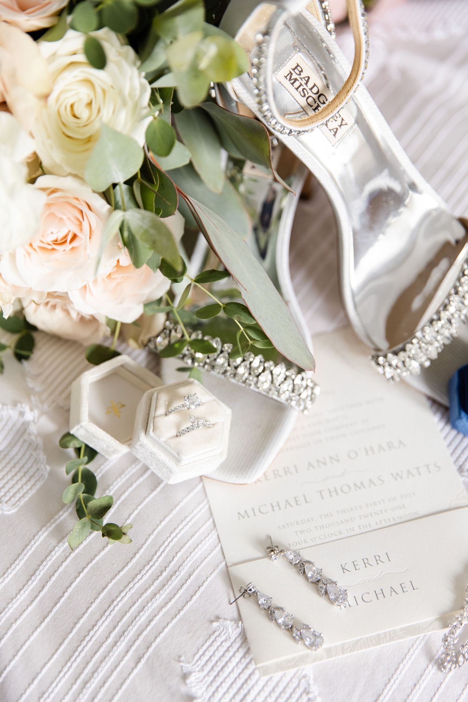 Wedding details with blush roses, silver shoes and invitation on white textured fabric.
