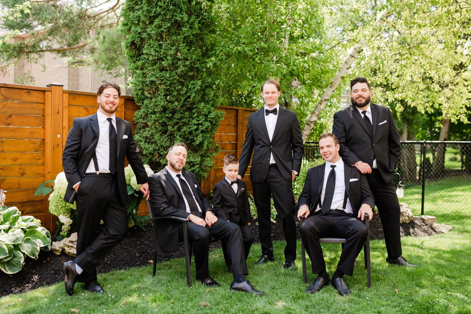 Groomsmen in formal black suits gathered for portrait in lush garden setting.
