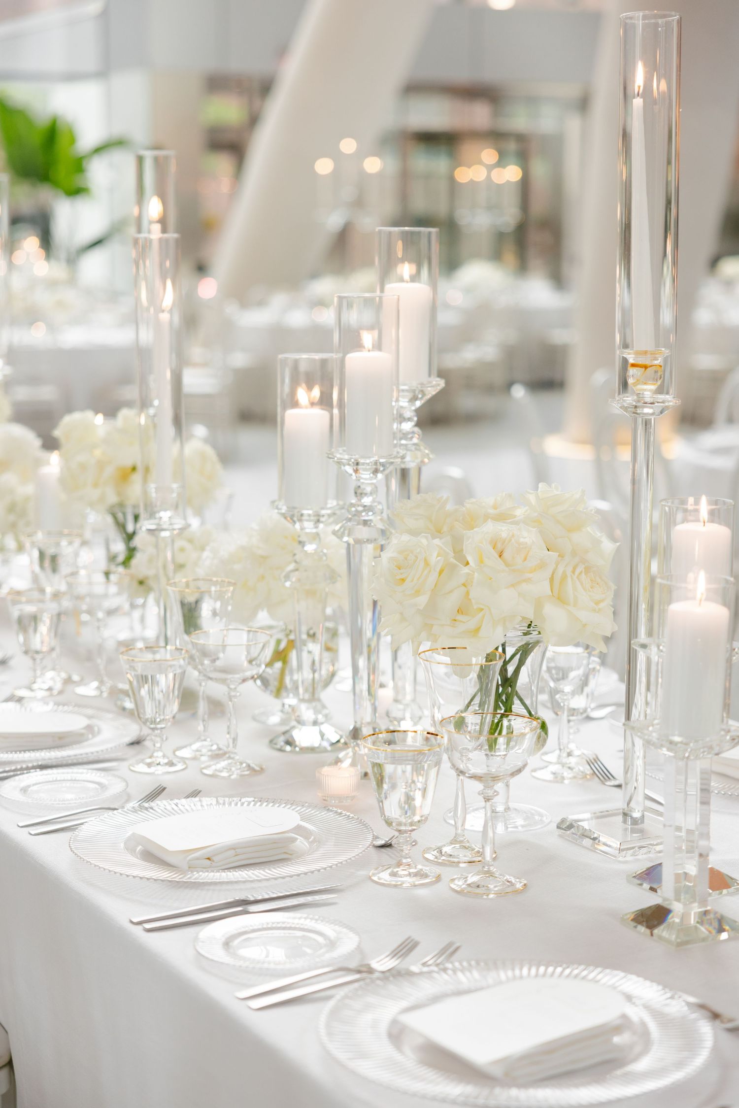 Elegant white wedding table setting with tall glass candleholders, white roses, and crystal glassware on crisp linens.
