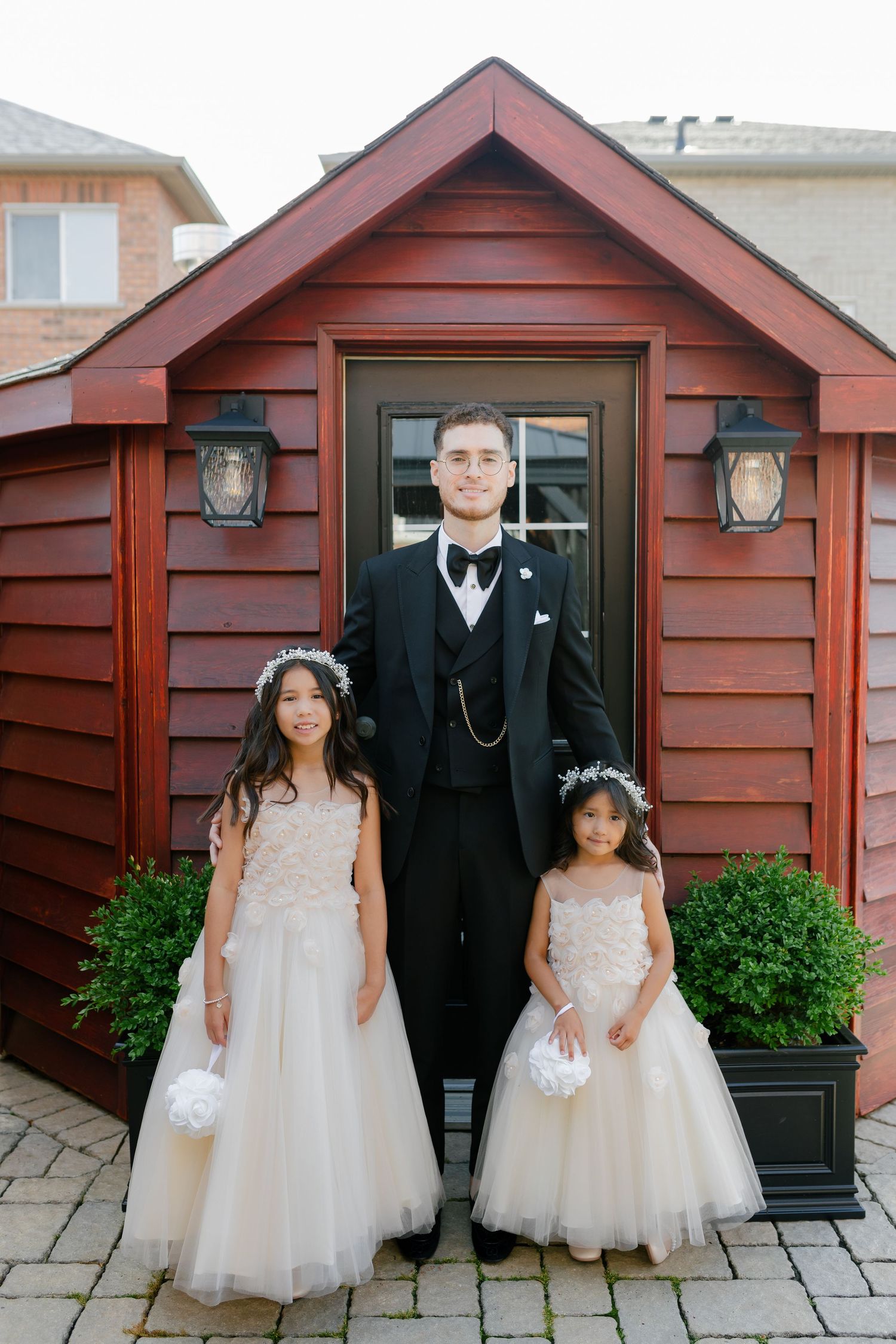 Two flower girls in white dresses pose with groomsman by red shed entrance.