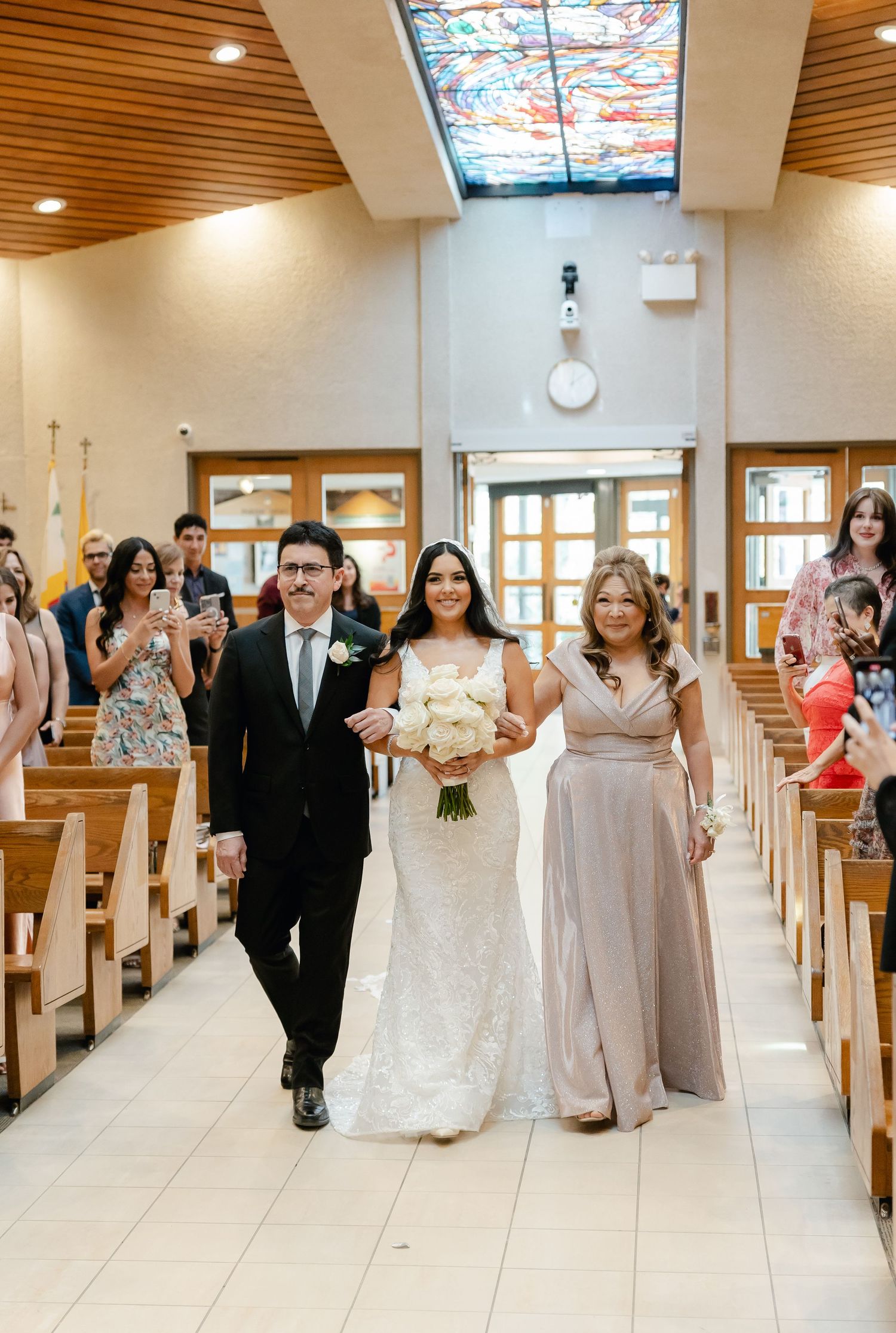 Wedding party walks down church aisle as guests look on beneath decorative skylight.