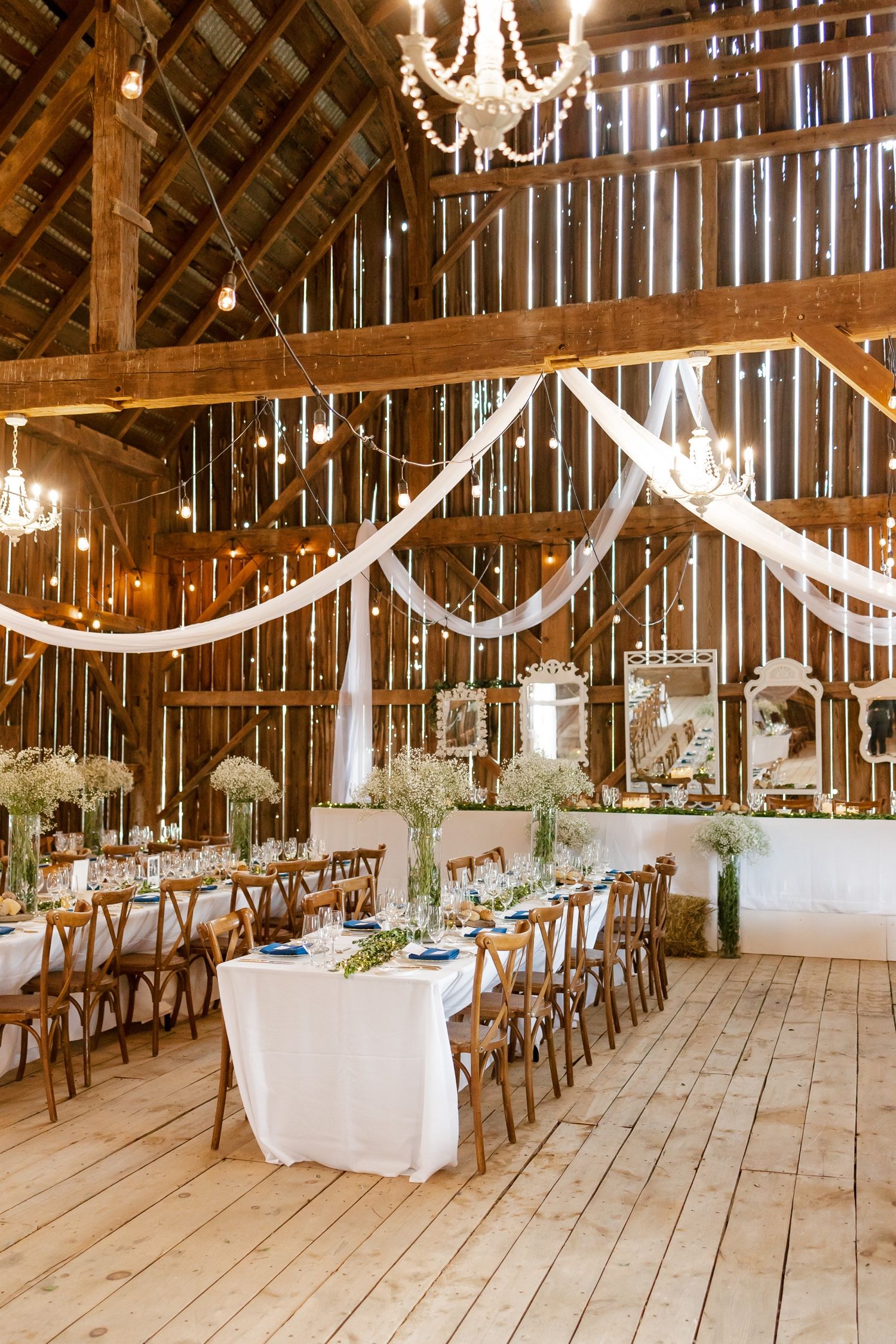 An elegantly decorated barn reception with chandeliers, white draping, and wooden cross-back chairs.