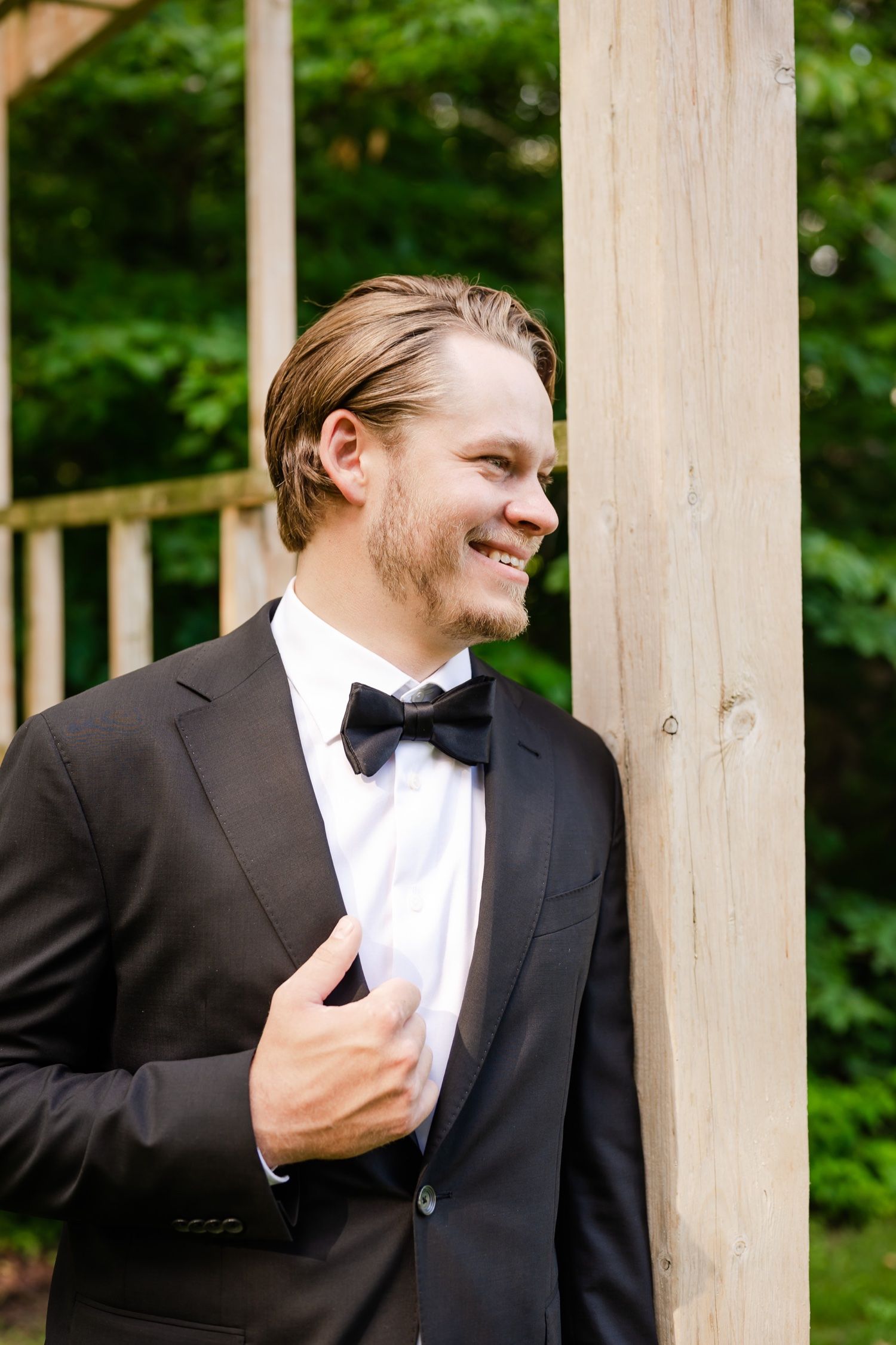 A groom in a classic black tuxedo and bow tie smiles while standing against a wooden pillar outdoors.