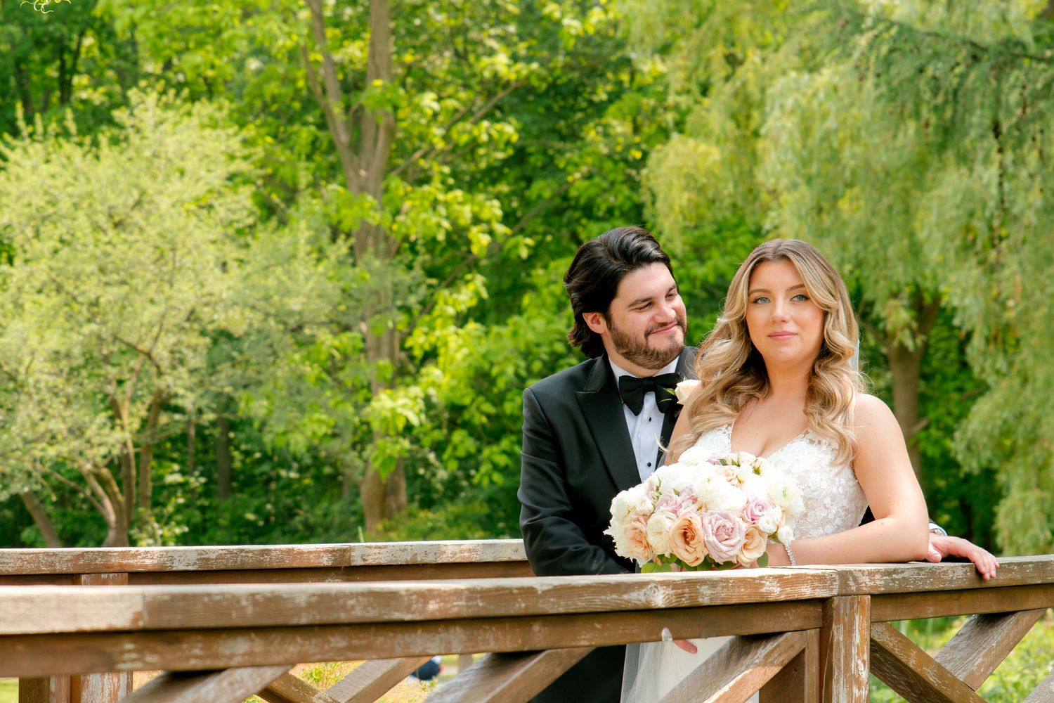Newlyweds pose on rustic wooden bridge surrounded by lush green foliage while holding pink bouquet.