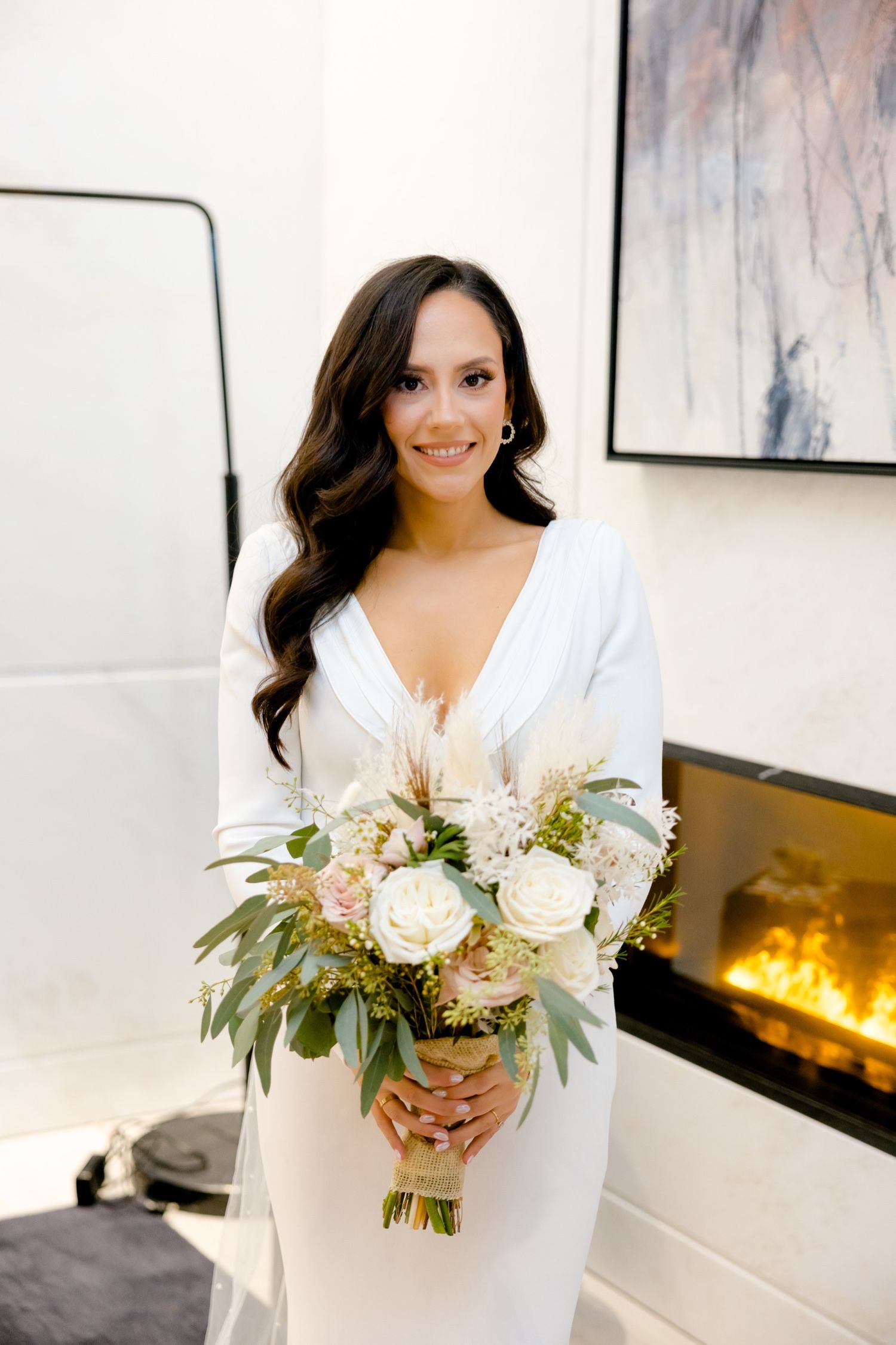 Bride in sleek white dress holds romantic bouquet of white roses and greenery by modern fireplace.