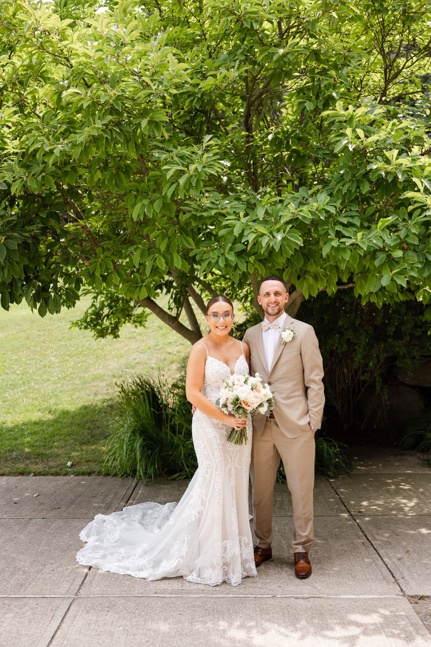 Wedding portrait of couple standing on concrete path beneath lush green tree.