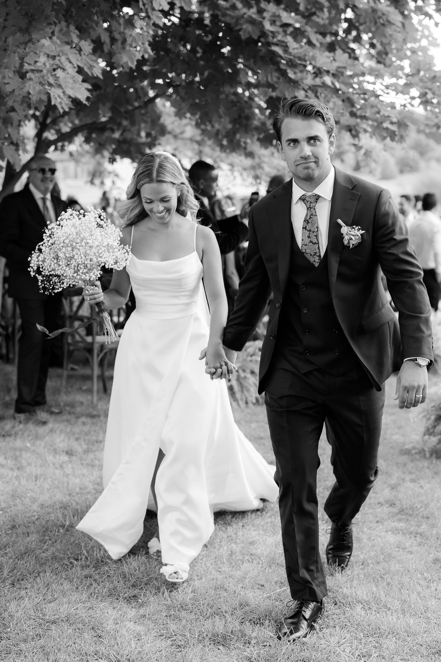 A black and white photo of a wedding couple walking outdoors with the bride holding baby's breath flowers.