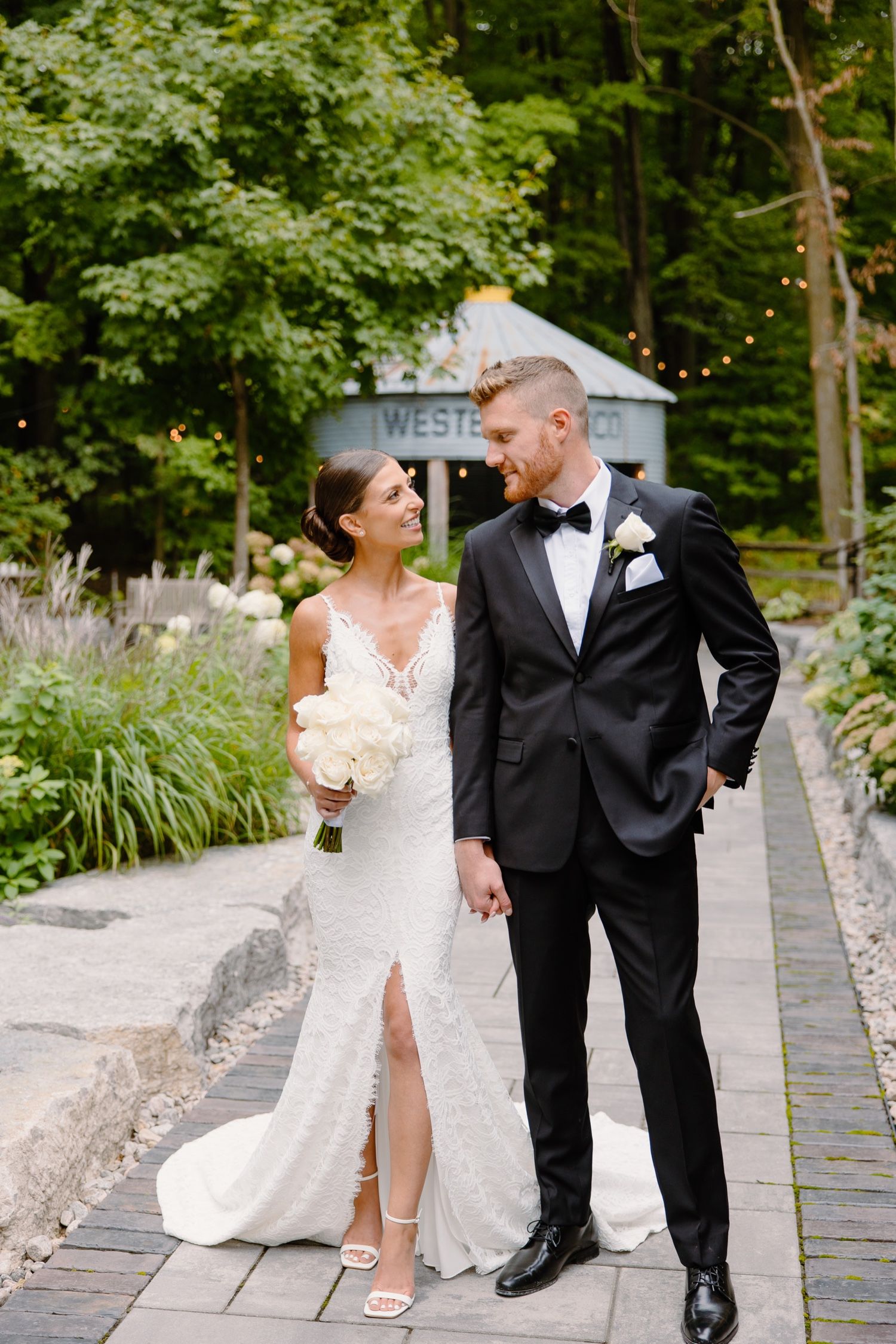 Wedding couple shares loving glance while posing on garden pathway near rustic venue.