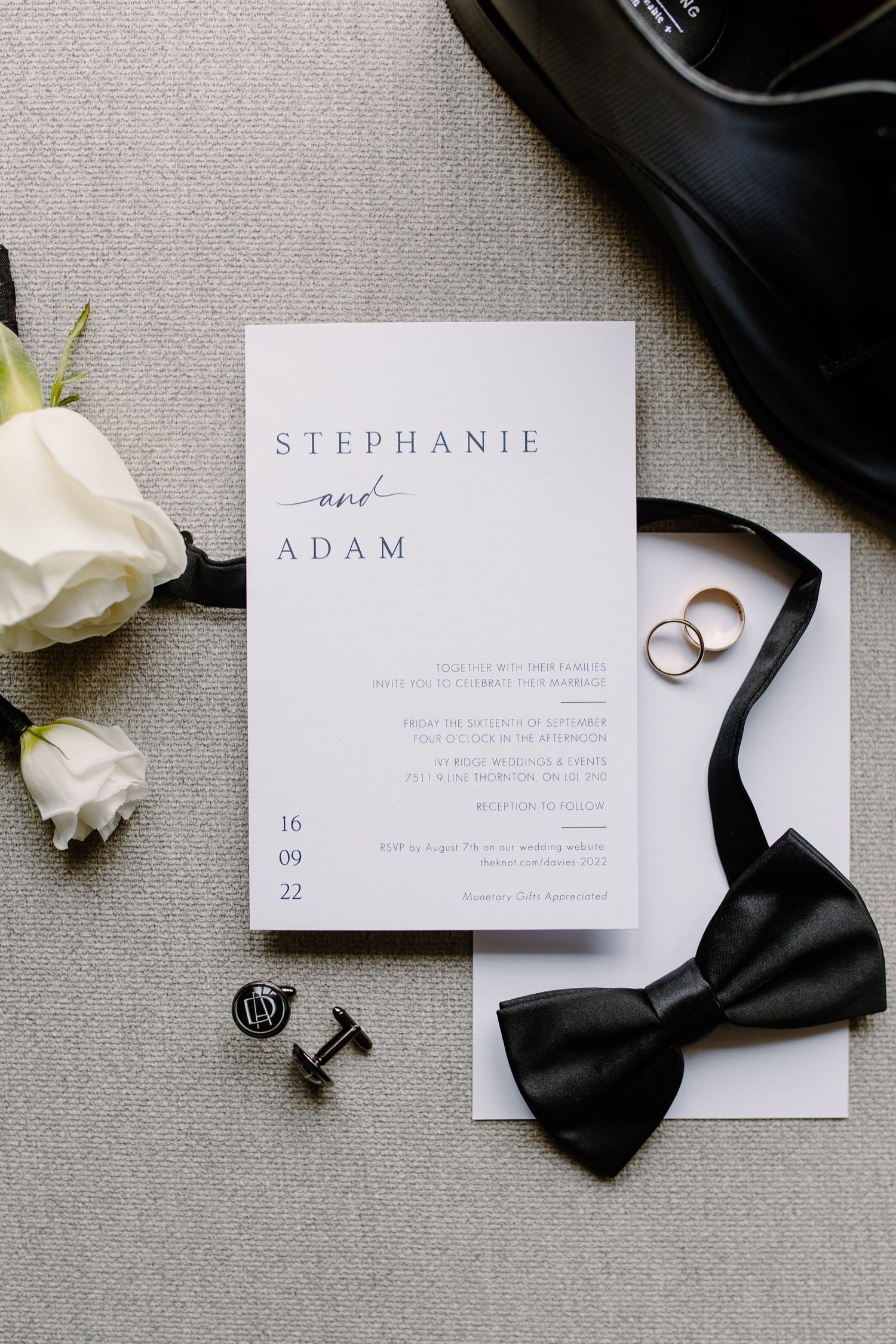Wedding invitation with black bow tie, rings and white roses on gray fabric background.
