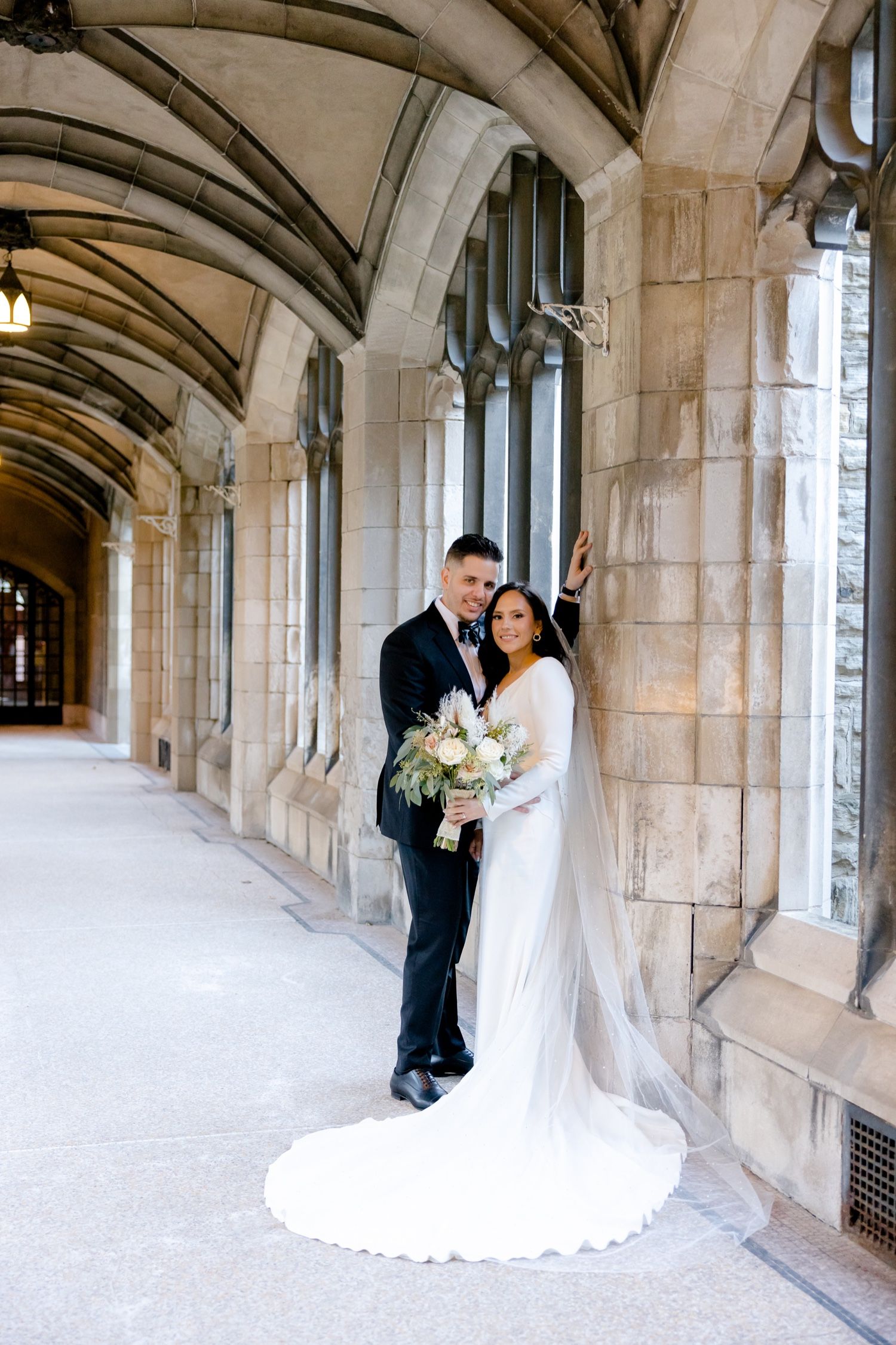 A newly married couple poses in a Gothic stone archway while holding a white and green bouquet.