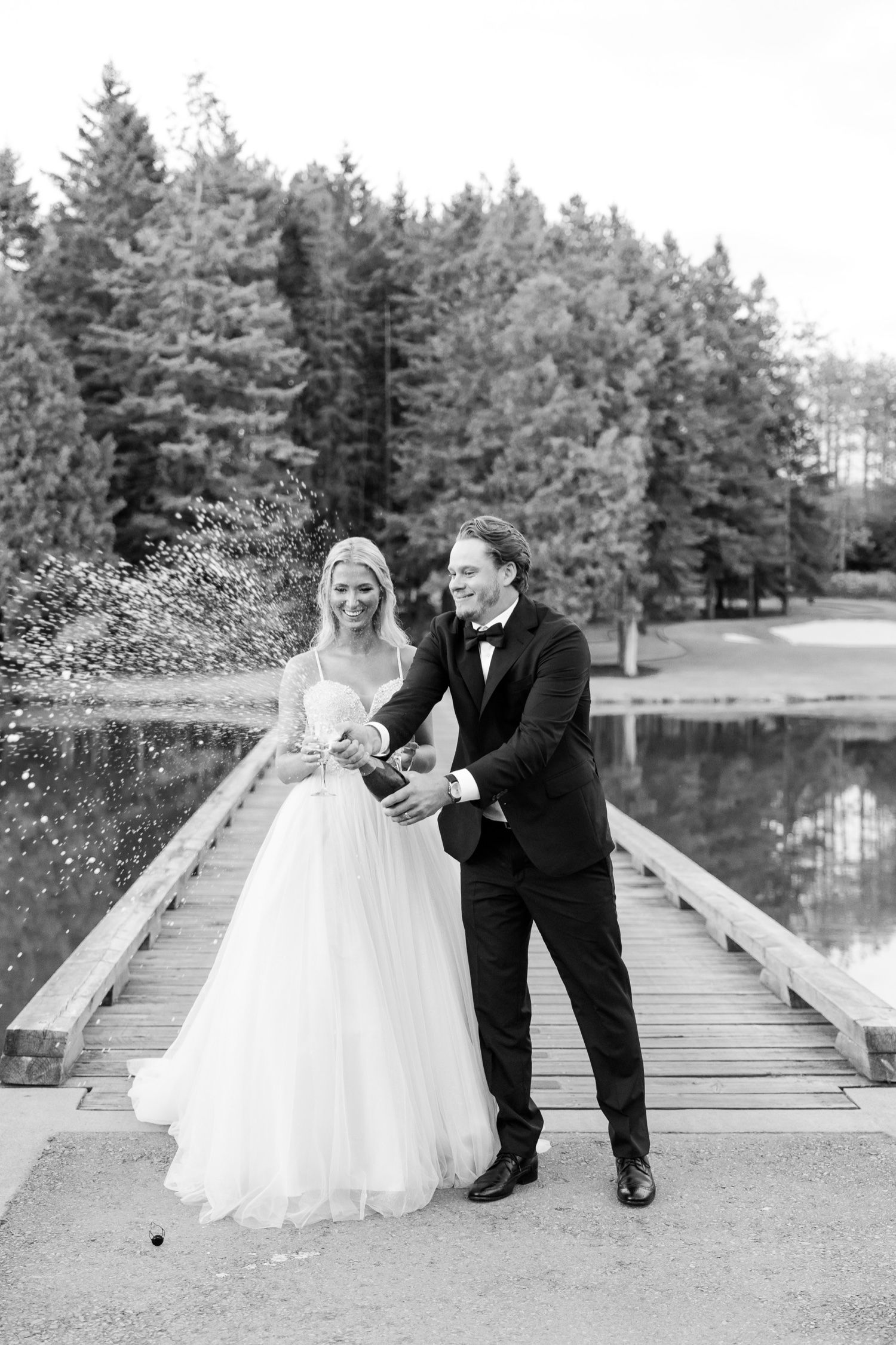 A black and white wedding photo of a couple celebrating with champagne on a wooden dock by a lake surrounded by evergreens.