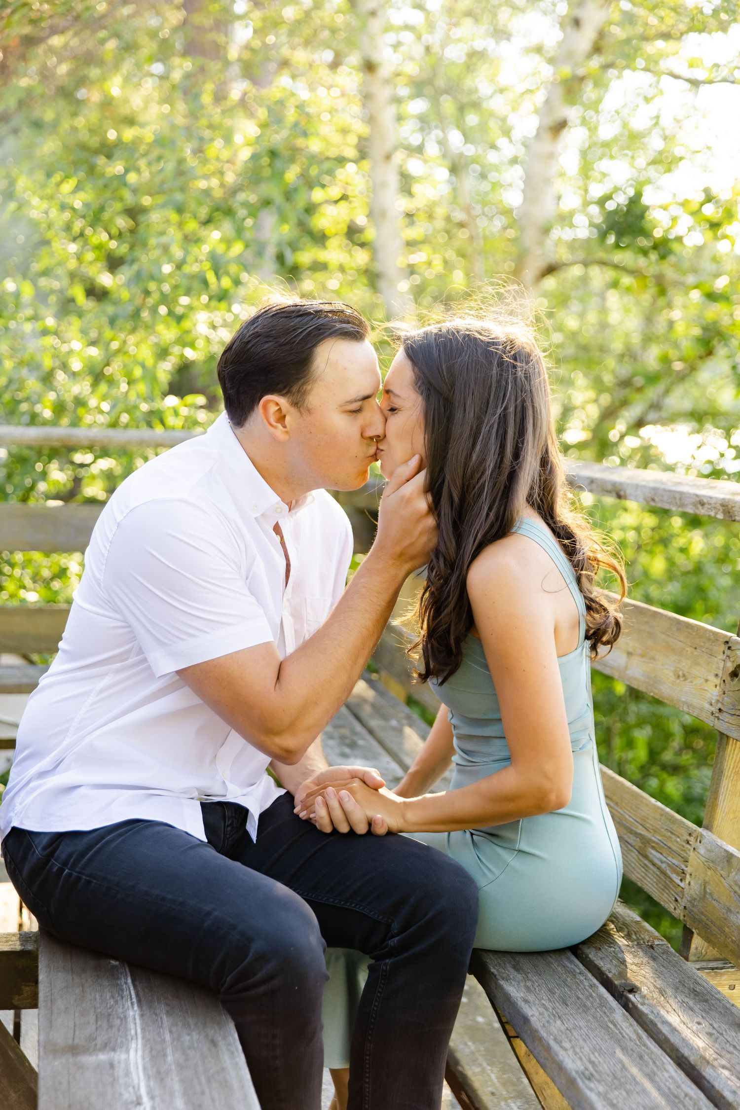 Couple shares romantic kiss while sitting on wooden bench in sunlit forest setting.