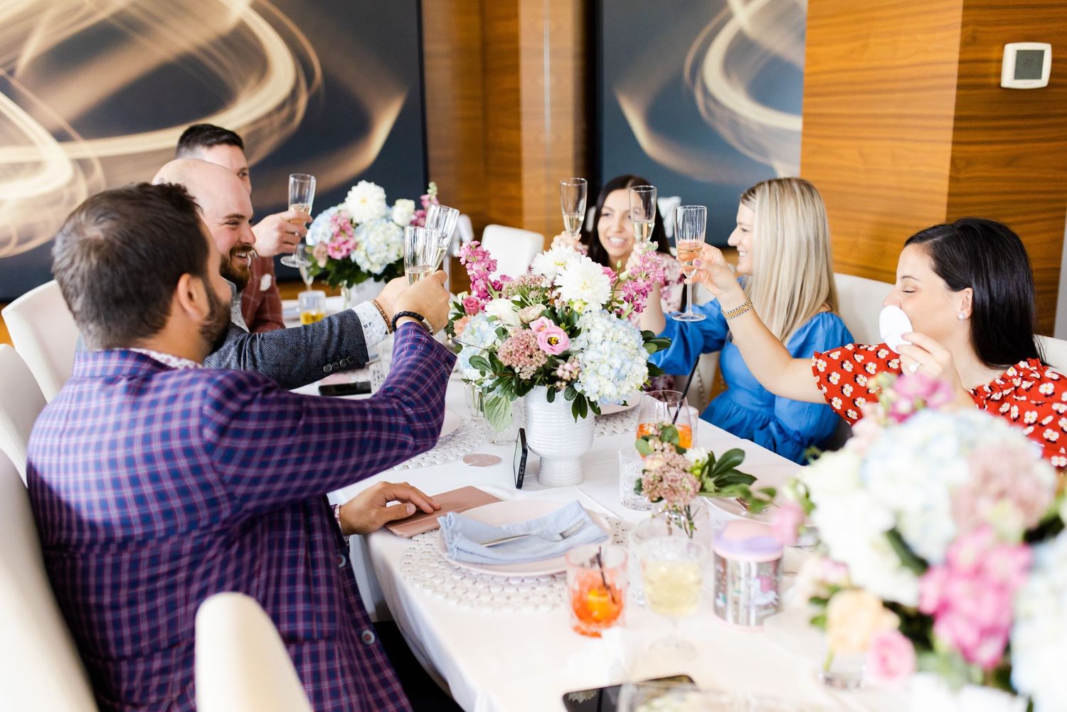 A festive dining table with guests toasting drinks and enjoying a meal with pink and white floral centerpieces.