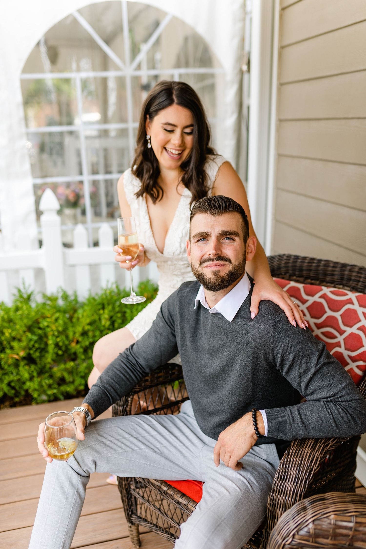 Couple enjoying drinks on a cozy porch with wicker furniture and decorative pillows.