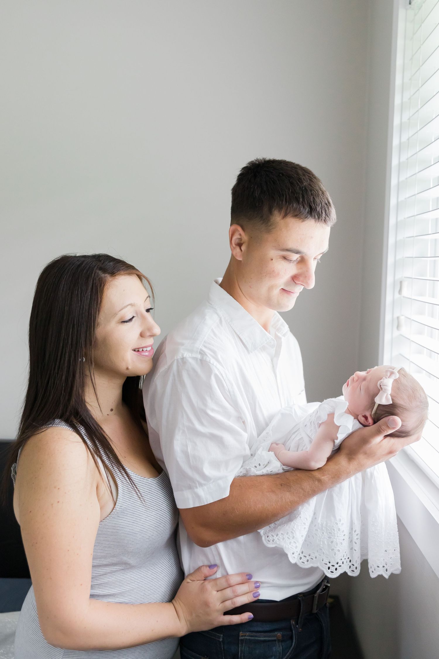 Parents admire their newborn baby together by window in minimalist setting.