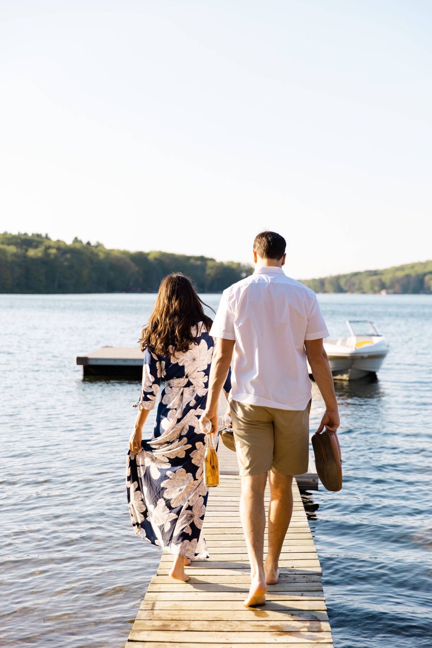Couple walking hand in hand on wooden pier with lake and forest in background.