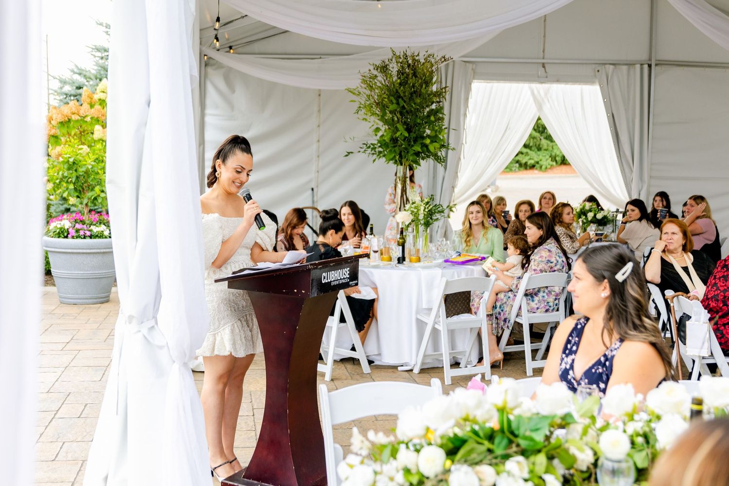 Speaker addresses guests at elegant outdoor reception under white tent with floral decor.