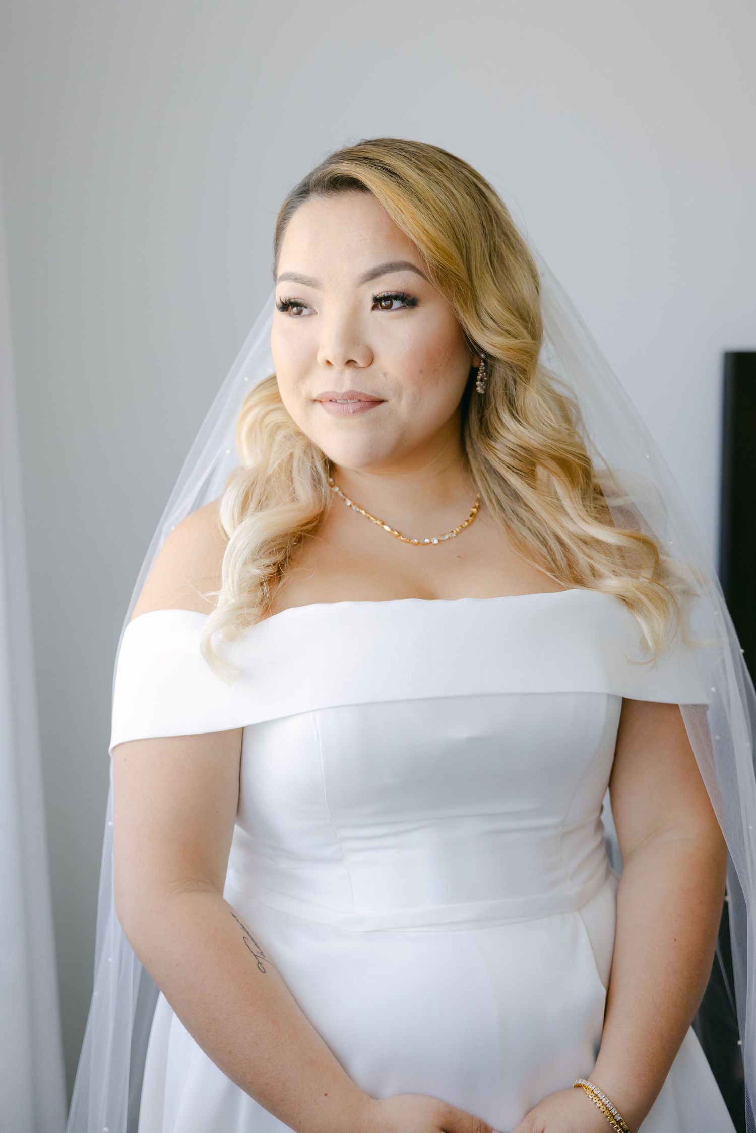 Bride wearing white off-shoulder gown and veil smiles while standing in natural light.