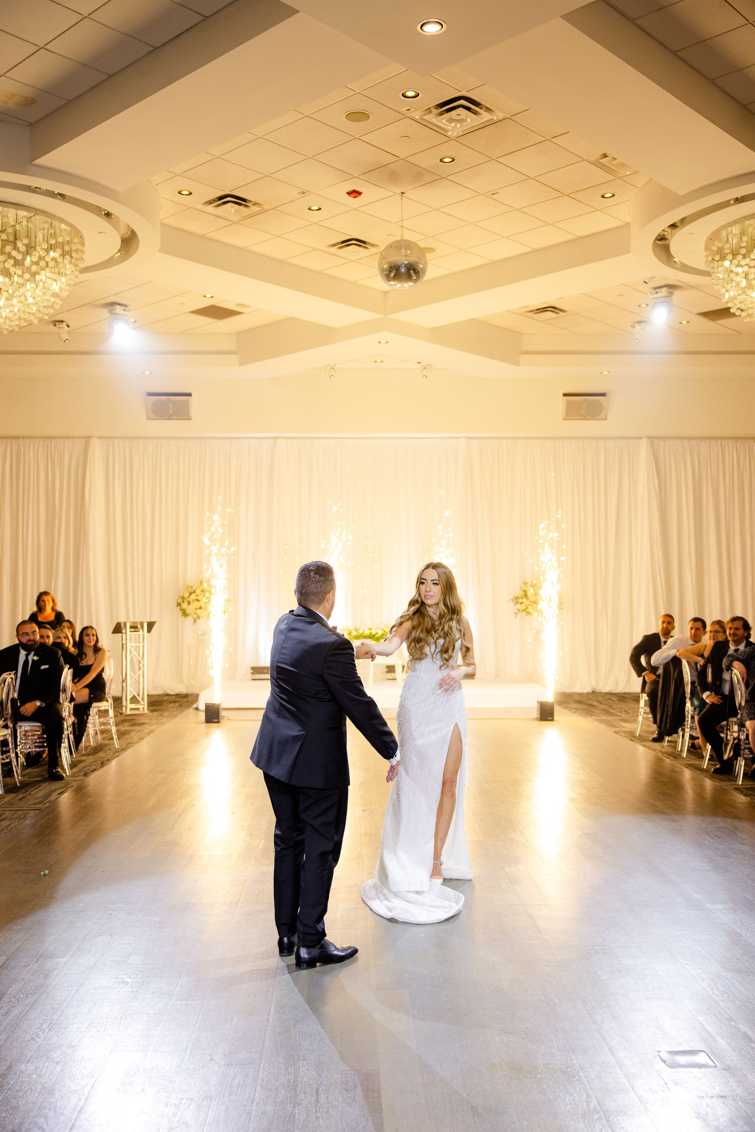 Wedding couple sharing their first dance in front of guests in an elegant ballroom with white decor.