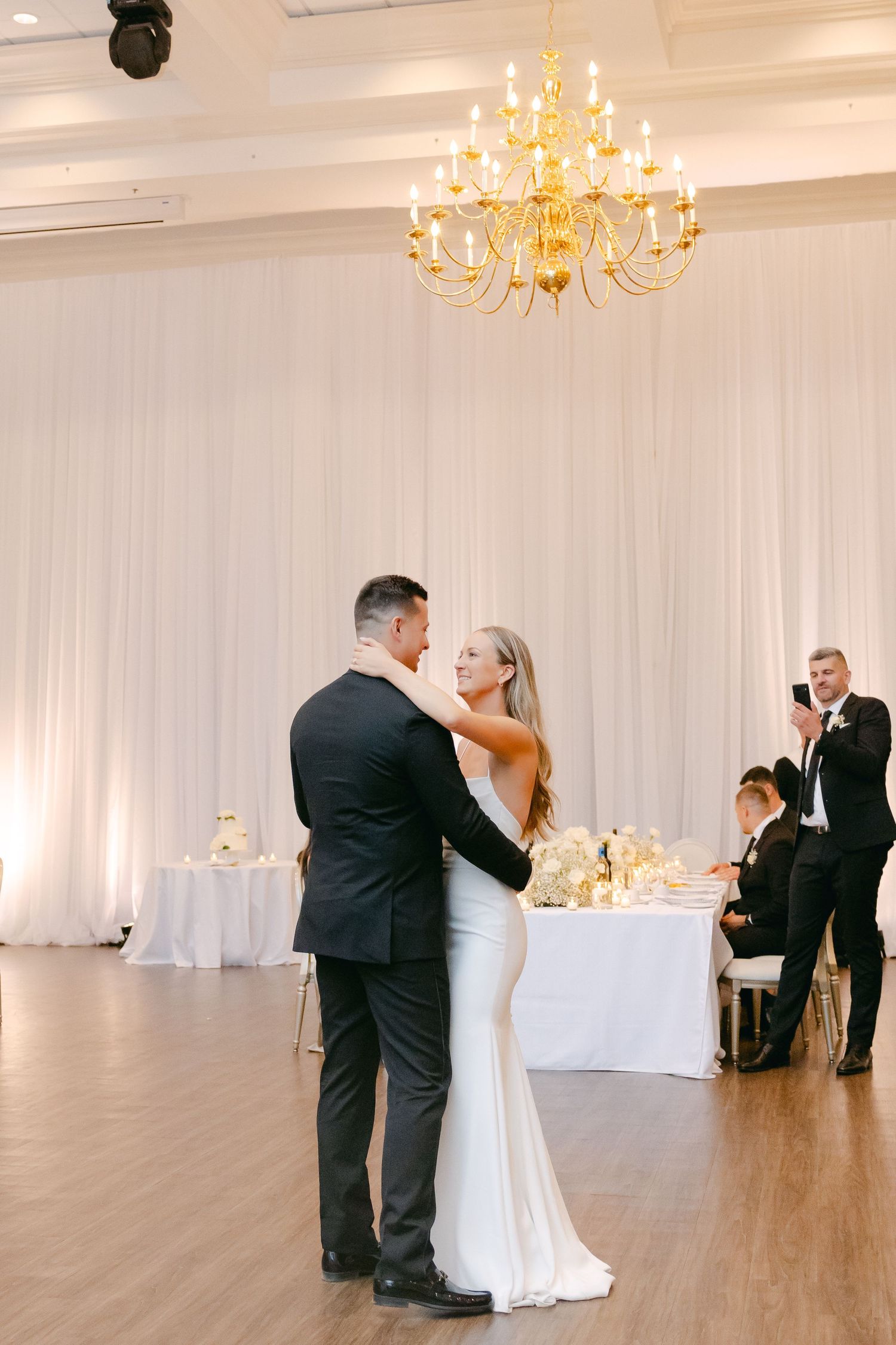 Intimate wedding dance moment beneath ornate golden chandelier in white-draped reception venue.