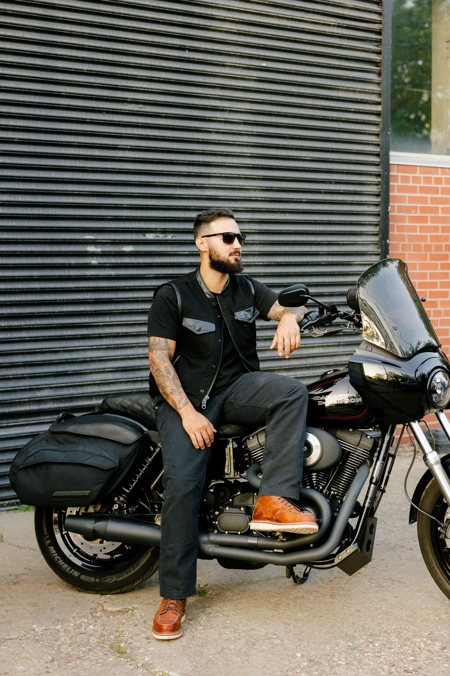 A motorcycle rider in black attire and sunglasses sits on a dark Harley Davidson bike against an industrial backdrop.