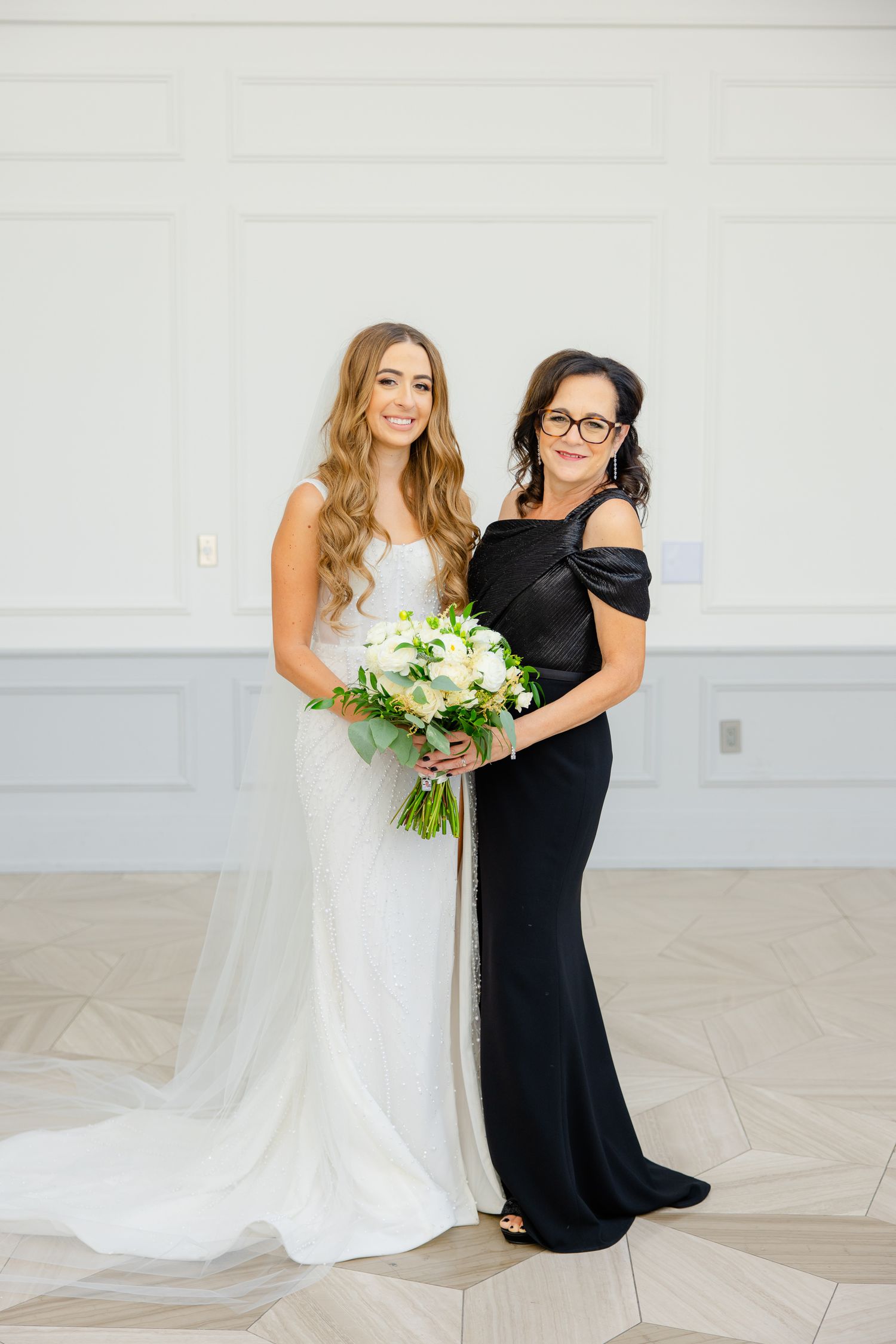 Elegant wedding portrait featuring two women in formal attire against a light gray wall.