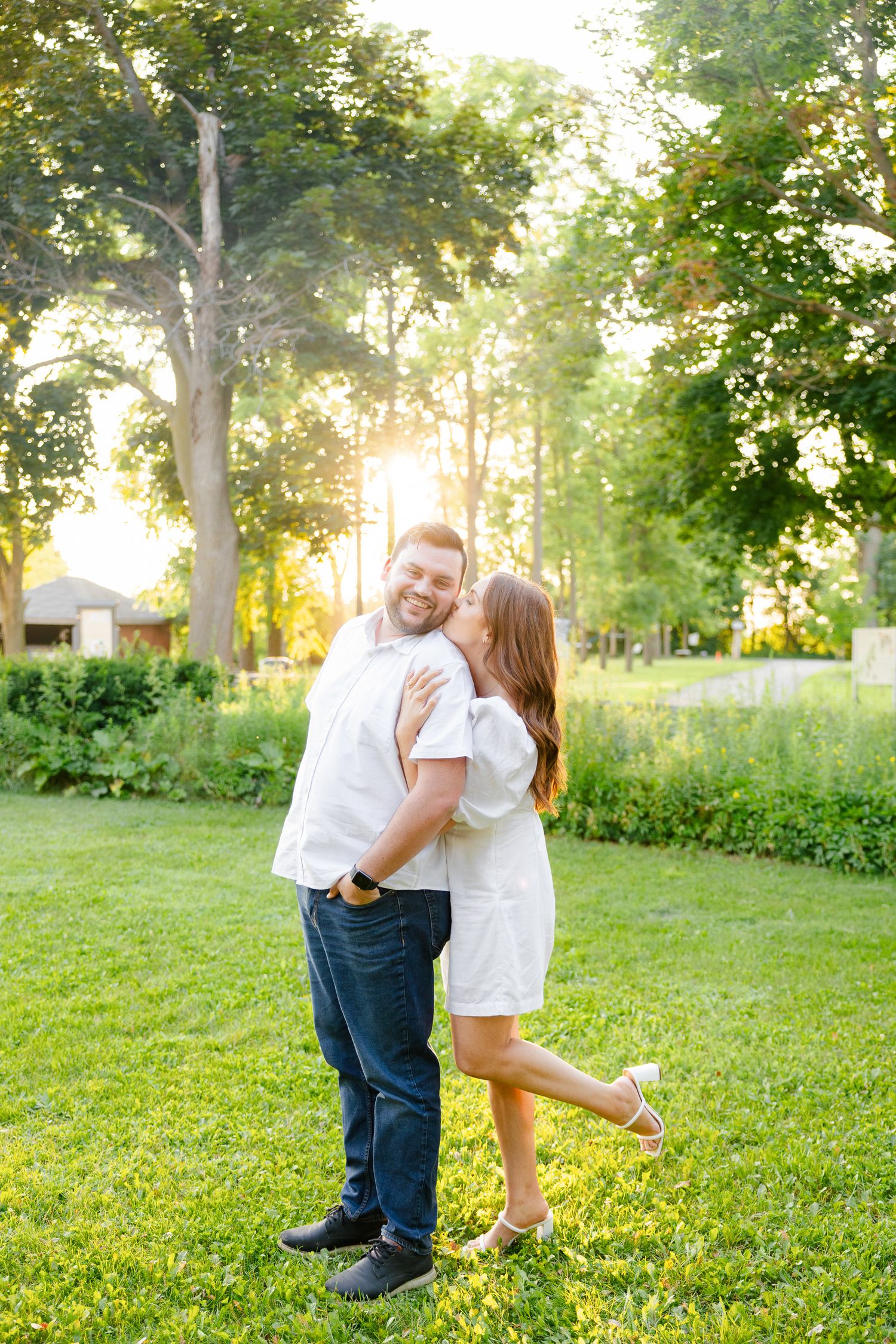 Playful couple in white outfits share romantic moment on sunny grass lawn.