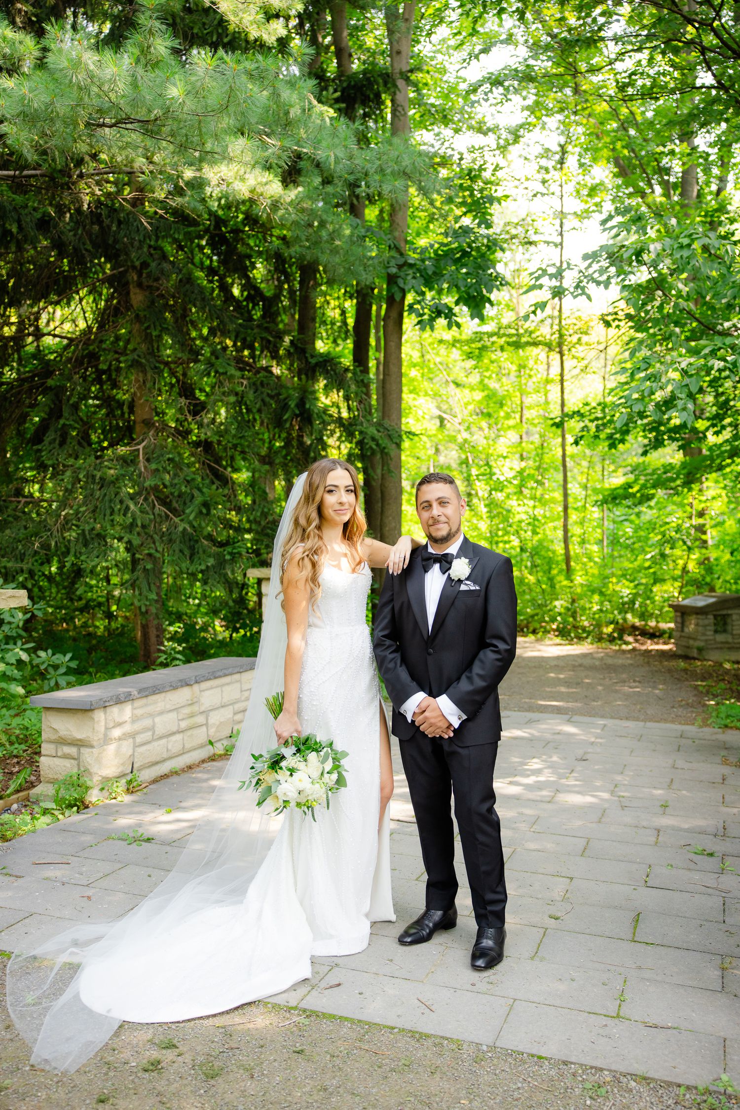 Elegant wedding couple poses on stone path with evergreen trees in background.