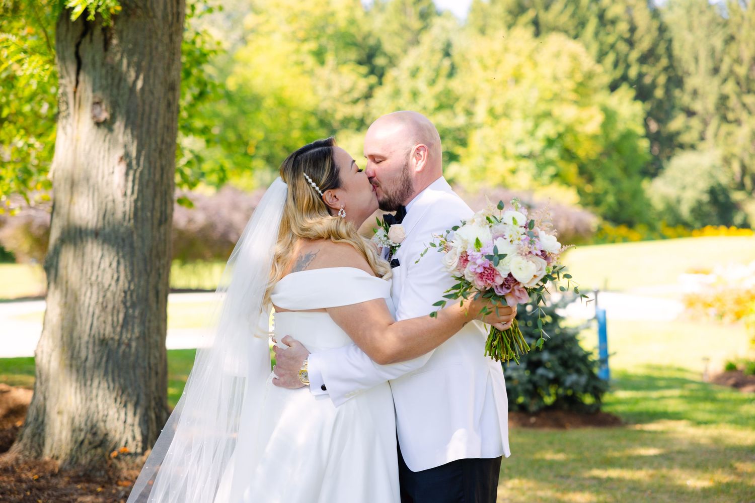 Tender wedding moment captured in a garden setting with sunlit trees in background.
