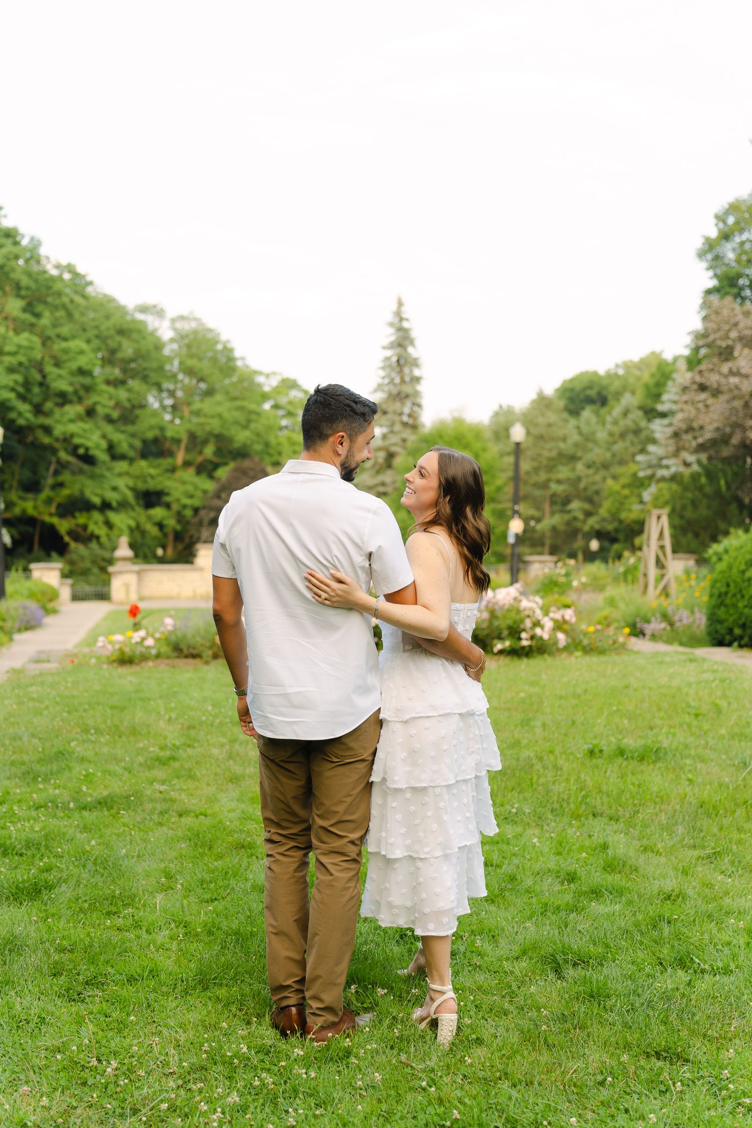 A couple shares a romantic moment while standing on grass with flowering bushes in the background.