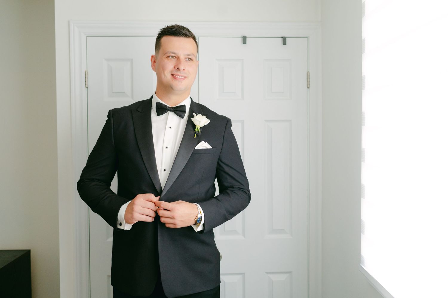 A groom smiles while adjusting his tuxedo jacket in a room with white doors.